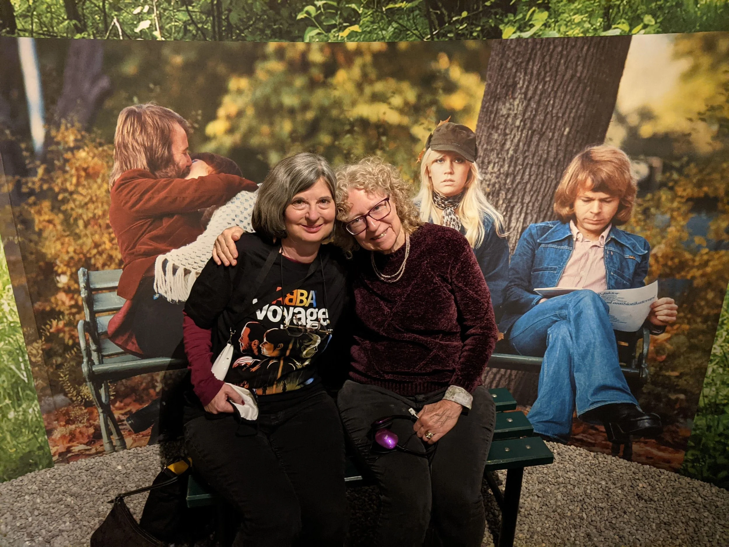 Dinah and Donna sit on a bench at ABBA: The Museum in Stockholm, posing in front of a backdrop of the Greatest Hits album cover featuring the band members.