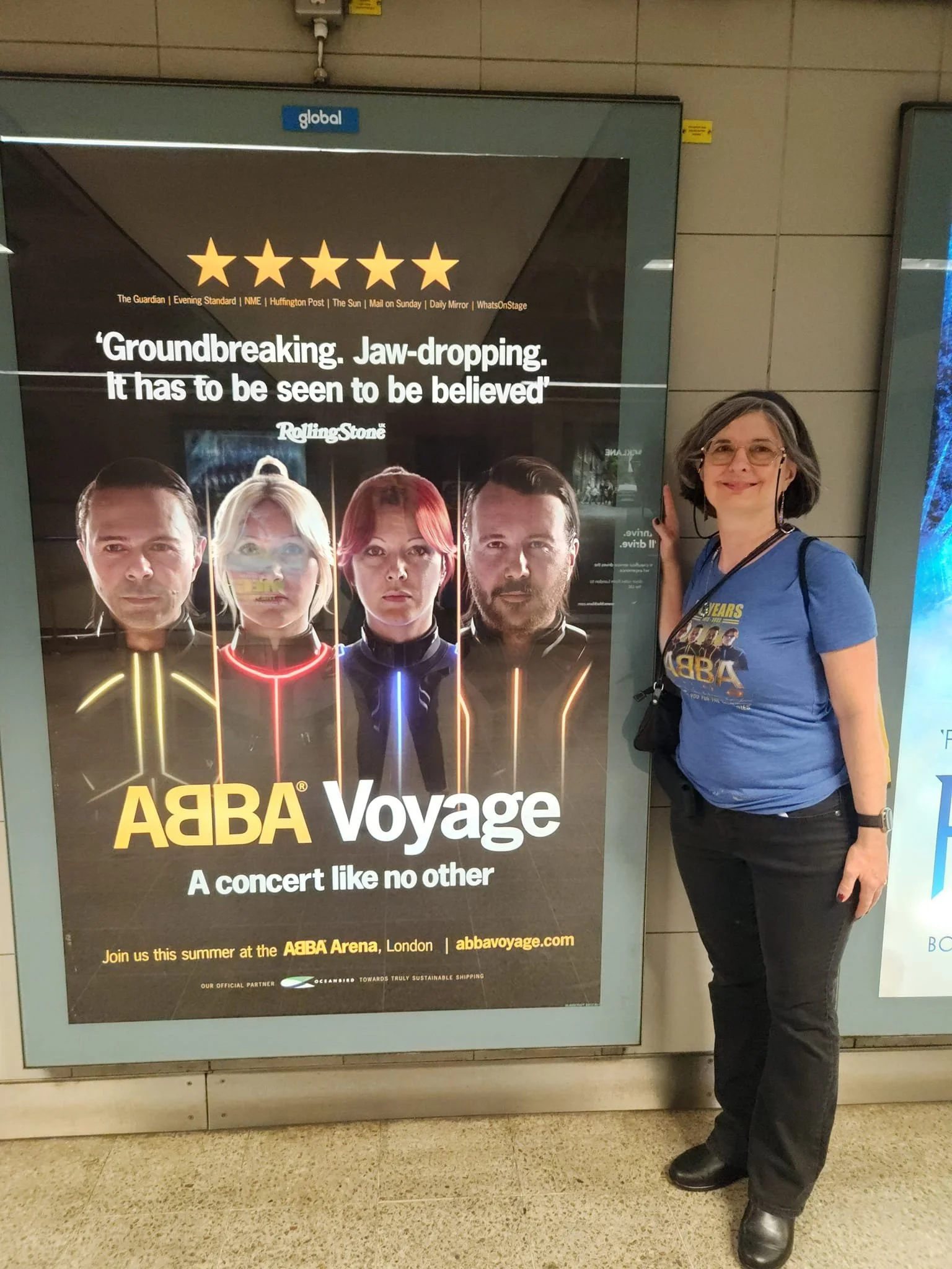 Dinah stands beside an ABBA Voyage concert poster in a London Underground station, featuring stylized portraits of the band and a five‑star review quote.