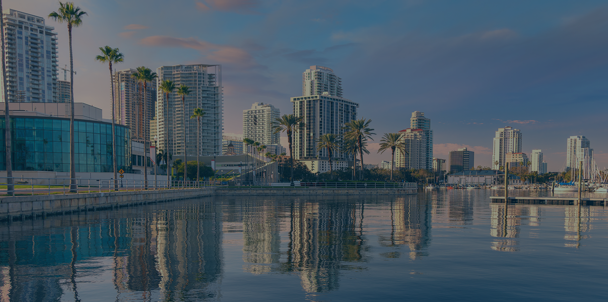 St. Petersburg, FL city skyline with modern high-rise buildings, palm trees, a waterfront, and boats docked in a marina at sunset.