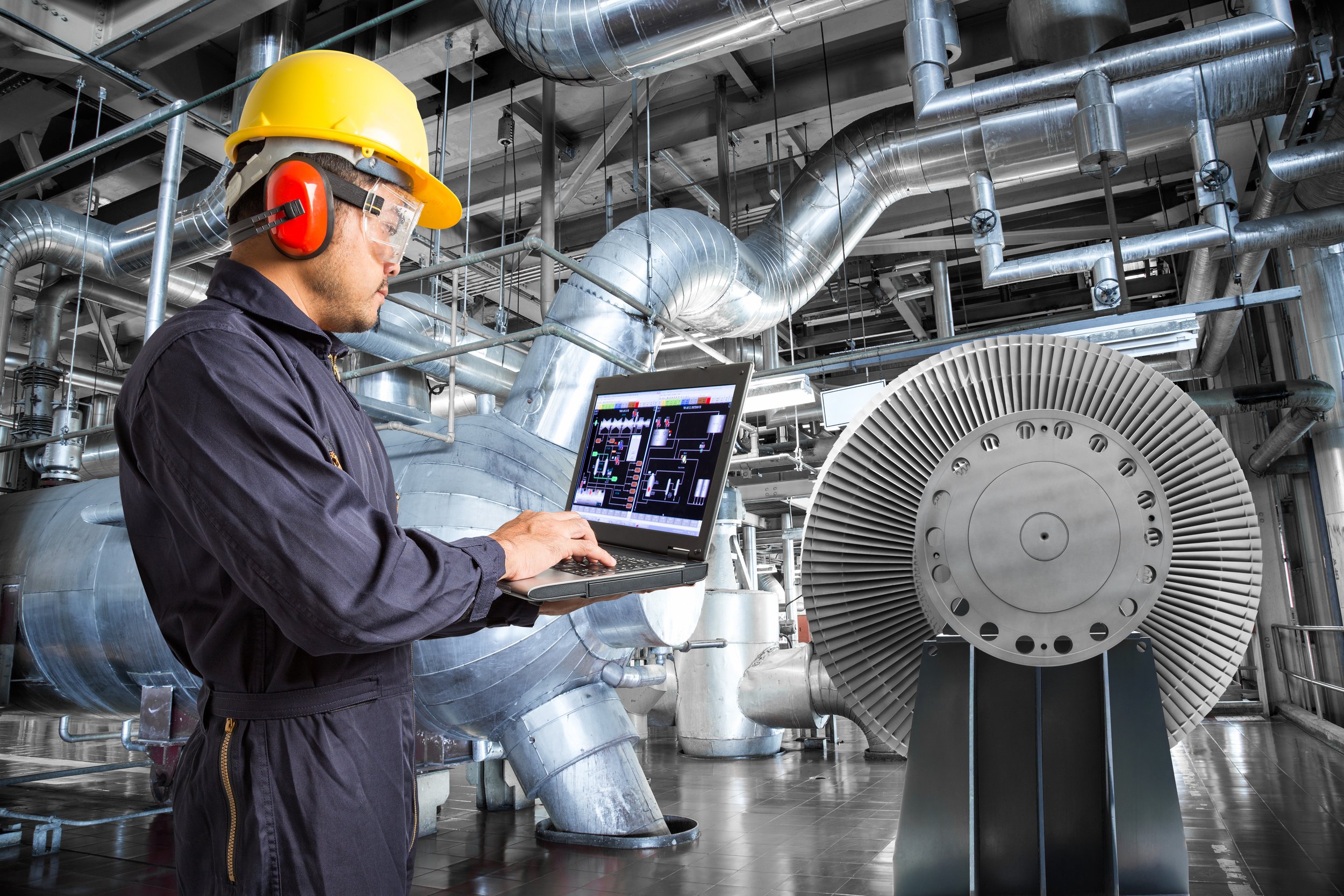 A worker in a yellow hard hat, safety glasses, and ear protection operating a laptop computer in an industrial facility filled with large metallic pipes and machinery.