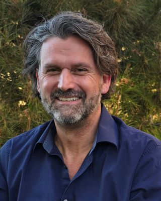 A smiling man with gray and brown hair and a beard, wearing a dark blue shirt, standing outdoors with trees in the background.
