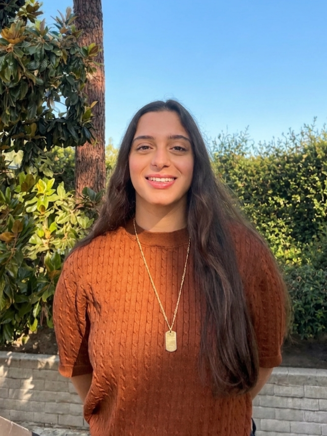 Smiling young woman with long dark hair, wearing a rust-colored cable-knit sweater and a necklace with a rectangular pendant, standing outdoors near trees and bushes on a sunny day.