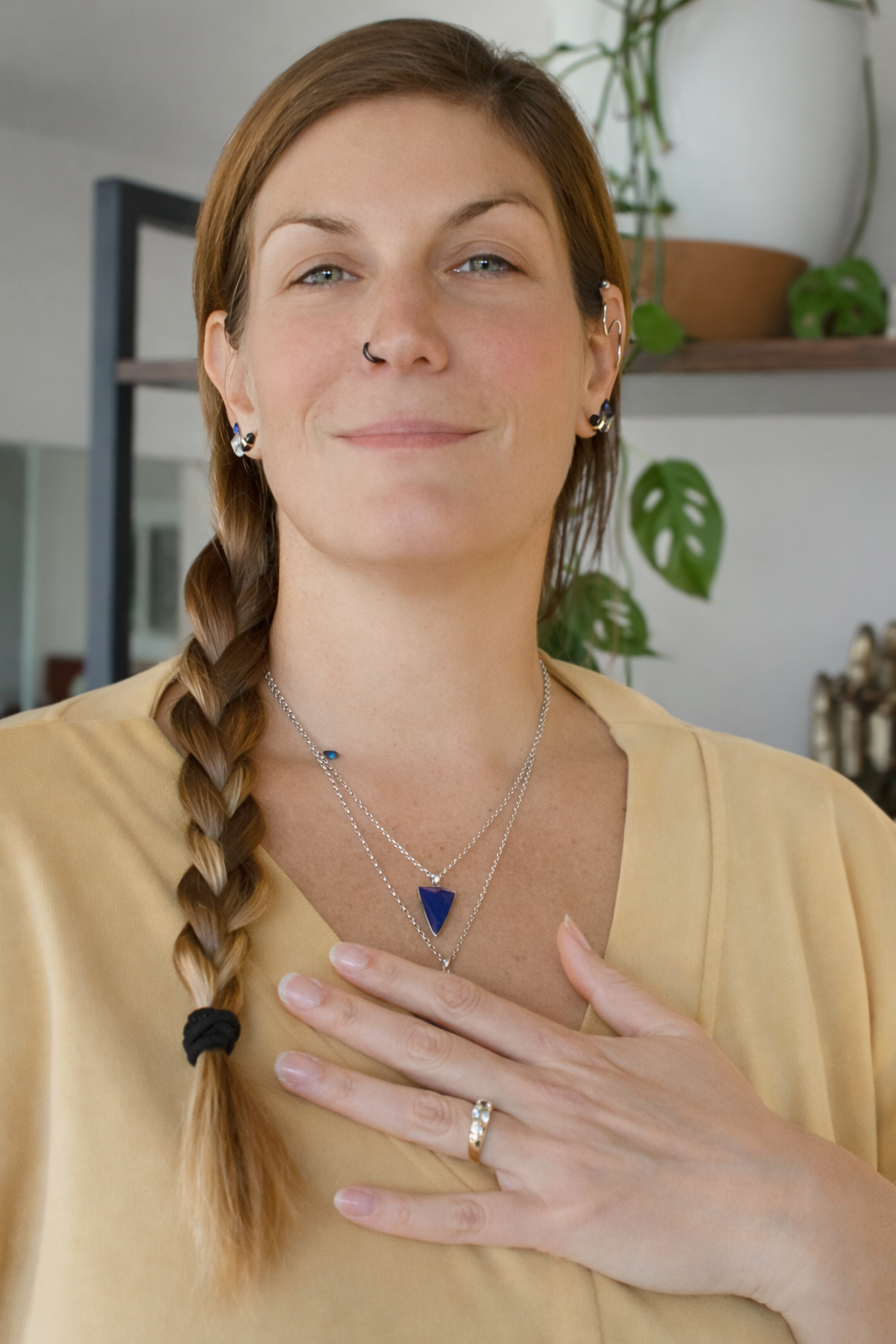 A woman with light skin and brown hair in a braid, wearing necklaces, rings, earrings, and a yellow top, standing indoors with plants and shelves in the background.