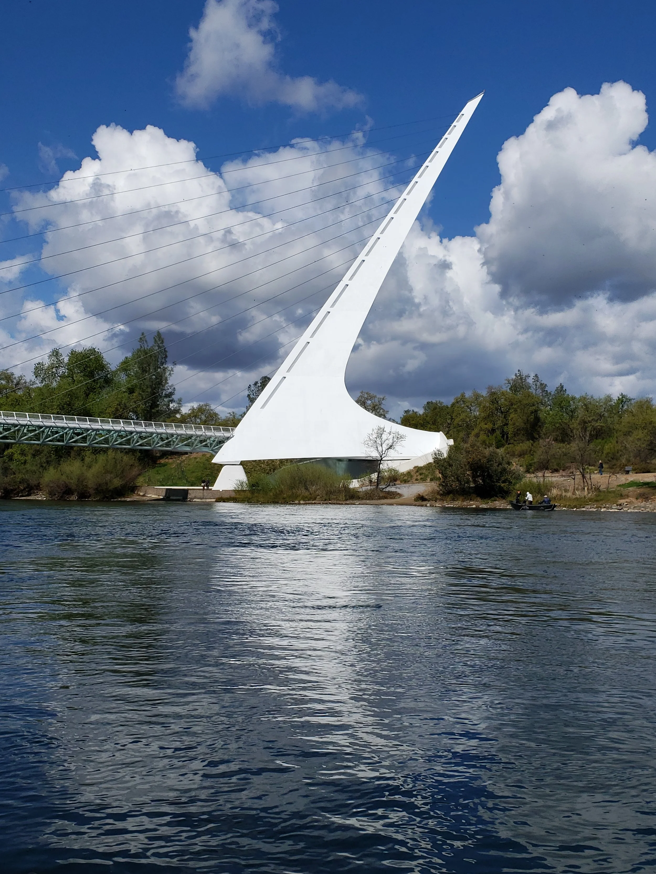 A bridge with a tall, white, pointed pylon and cable supports, spanning over a river with calm water and a partly cloudy sky in the background.