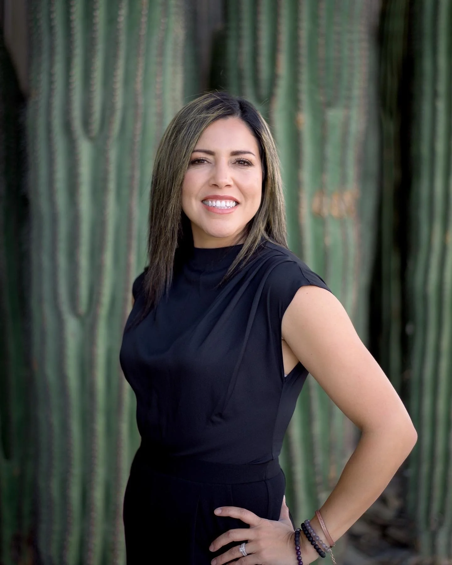 A woman with light skin and dark brown hair with blonde highlights, wearing a black sleeveless top and multiple bracelets, stands in front of tall cacti, smiling with her hand on her hip.