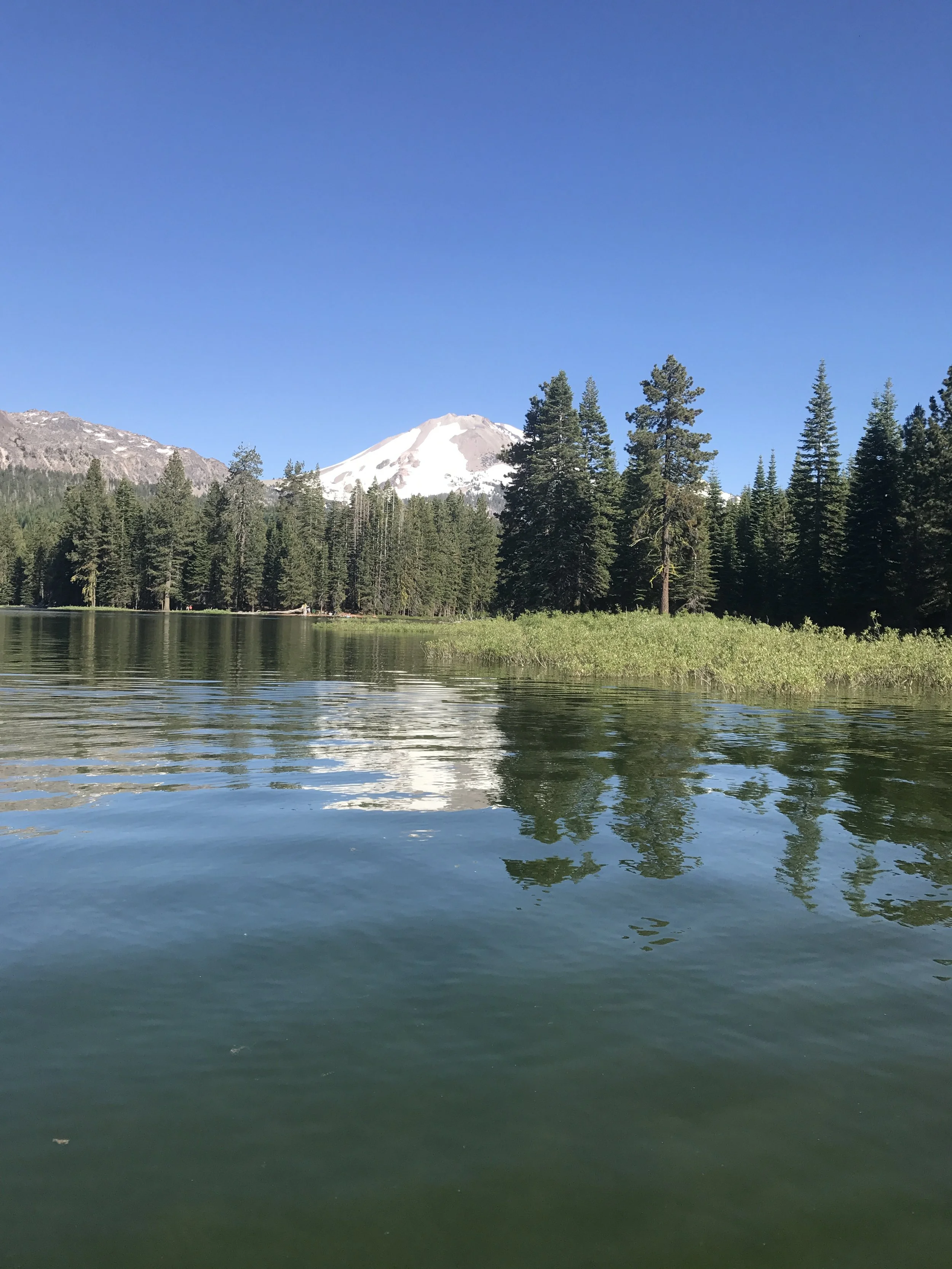 A lake with calm water reflecting green trees and mountains, including a snow-capped peak, under a clear blue sky.