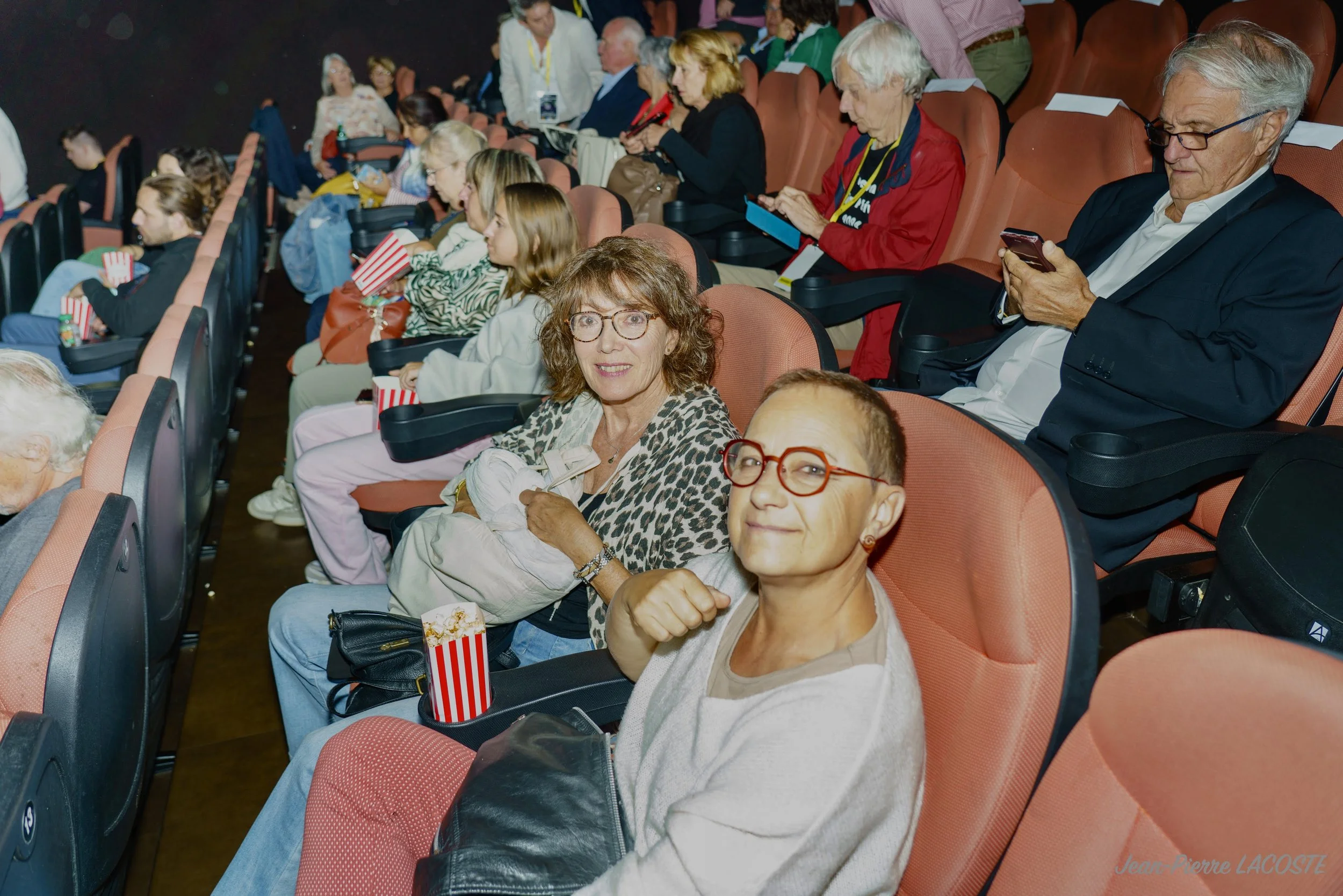 Groupe de personnes assises dans une salle de cinéma, certaines utilisant leurs téléphones et mangeant du popcorn.