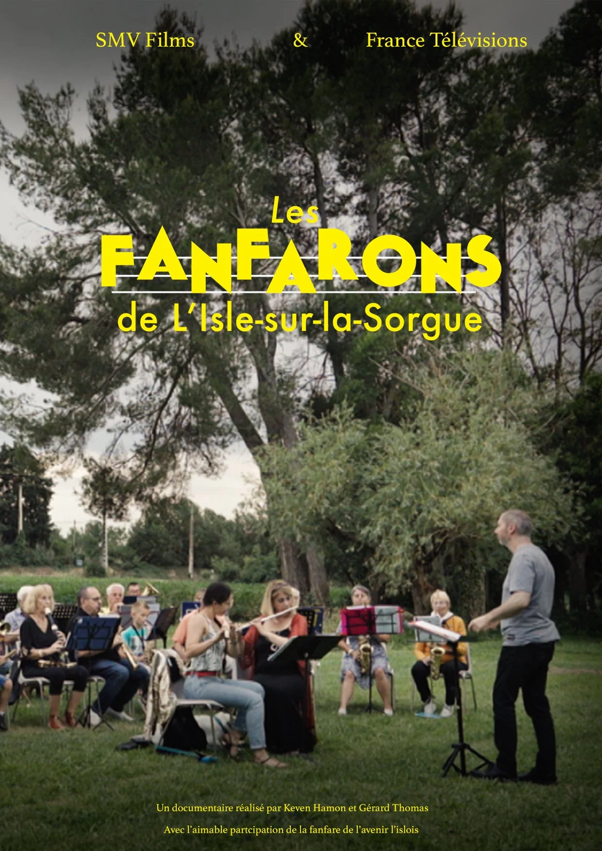 Un groupe d'instruments à vent, tels que des flûtes, clarinettes, saxophones et trompettes, jouant dans un parc lors d'un concert de musique. Un chef d'orchestre dirige le groupe, situé devant eux, avec un fond d'arbres et un ciel nuageux. Le texte e