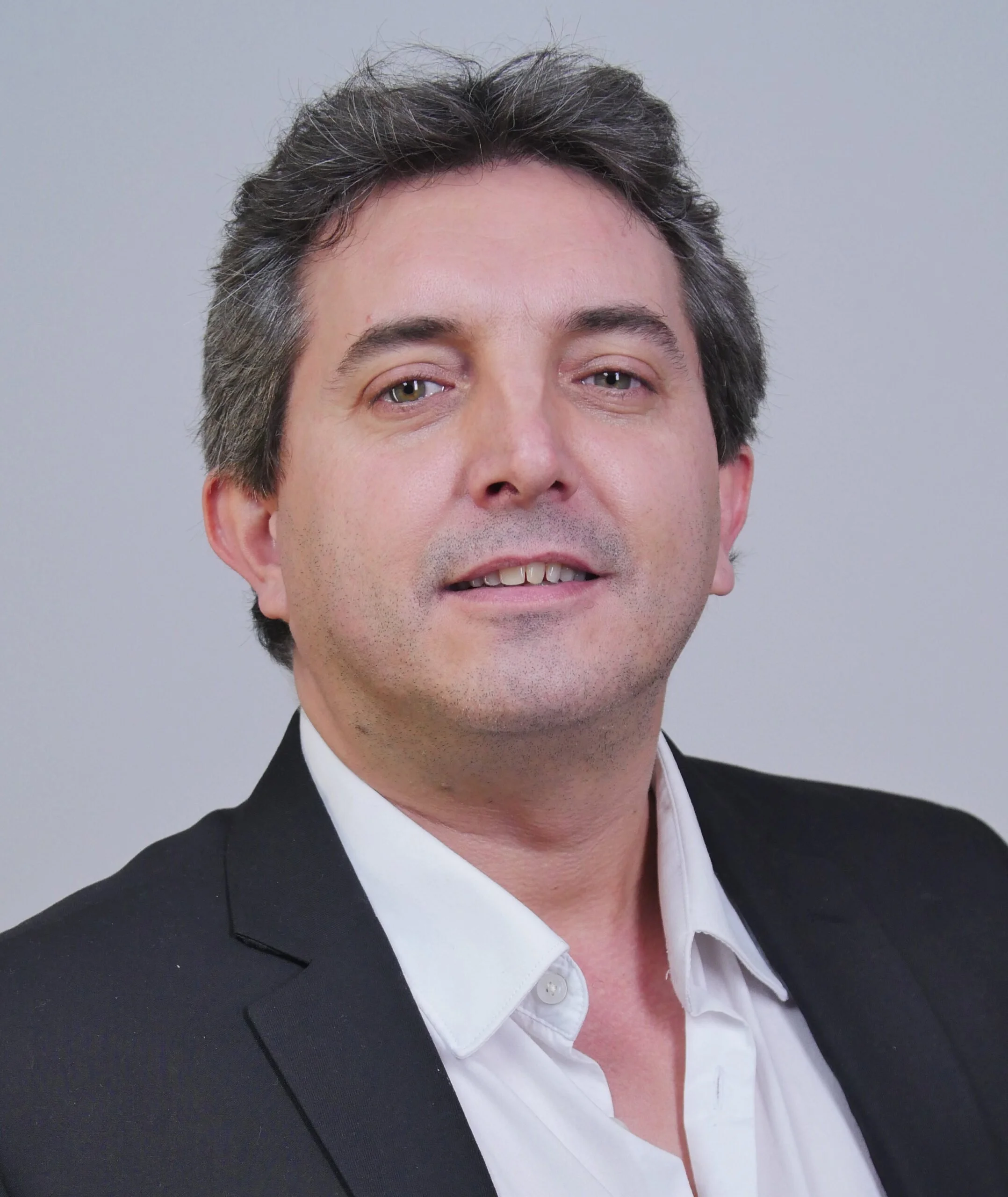 Photo d'un homme portant un costume noir et une chemise blanche, souriant légèrement, avec des cheveux bruns et courts.