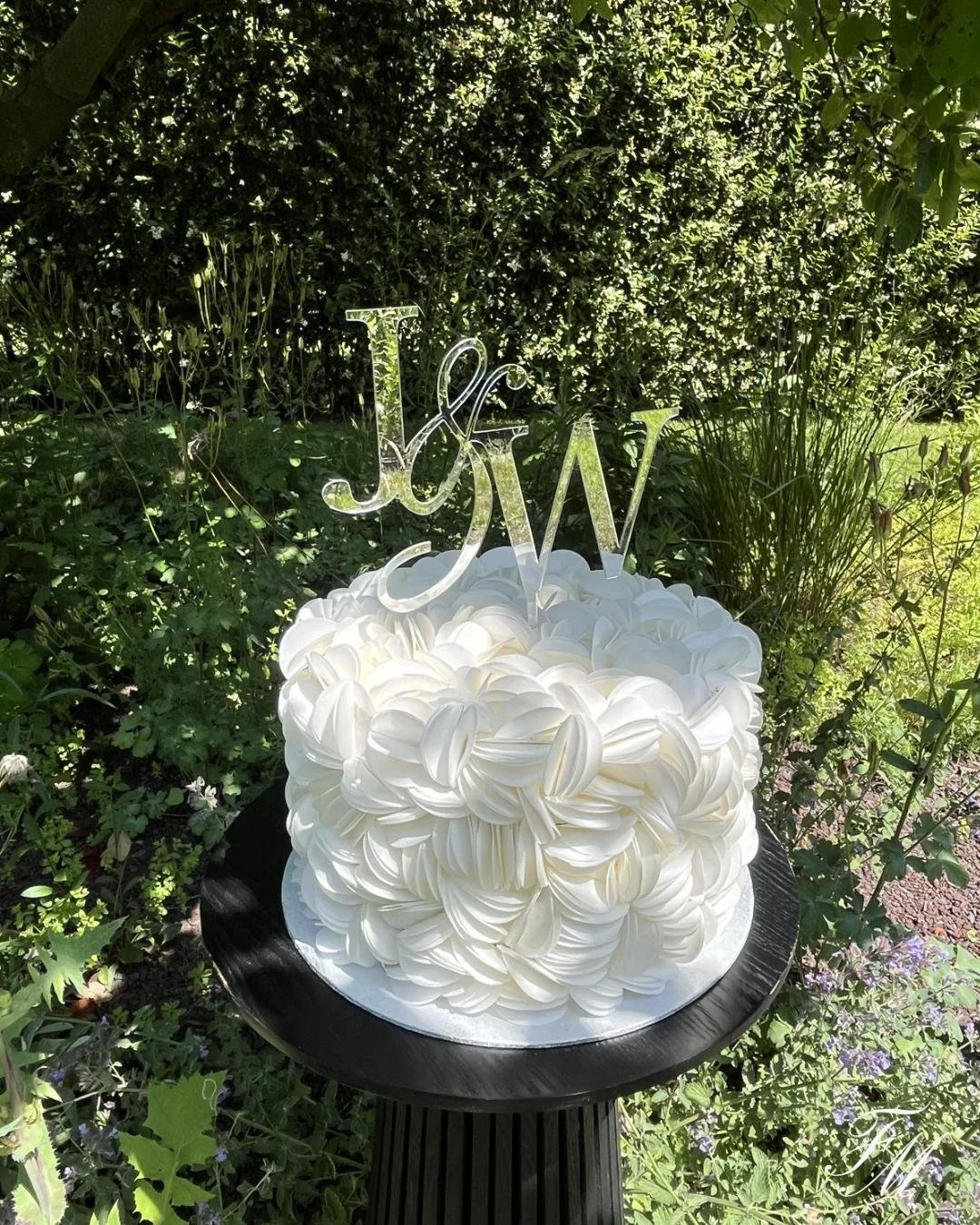 Een wit bruidstaart met een decoratie van krullen, geplaatst op een zwart voetstuk in een tuin, met een glitterend fotolabel met de initialen J&W.