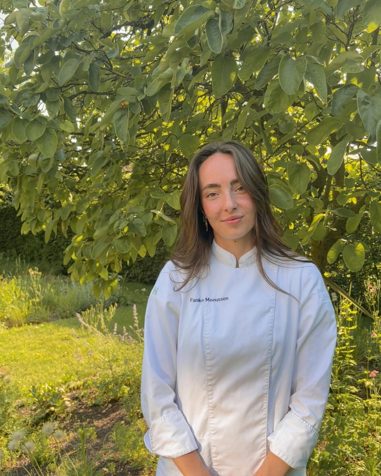 Vrouw in een witte chef-kok uniform met de naam 'Famke Meeussen' op de borst, staat buiten voor een grote boom met groene bladeren, zonnige tuin in de achtergrond.