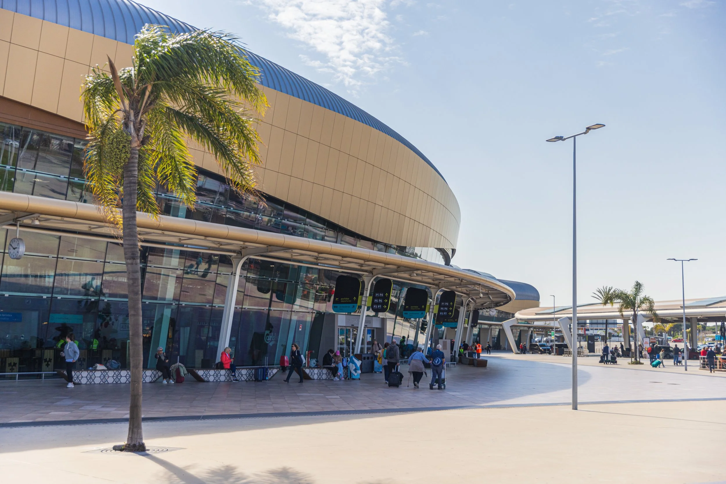 People walking and sitting outside a modern transportation hub with a curved architectural design, palm trees, and clear blue sky.