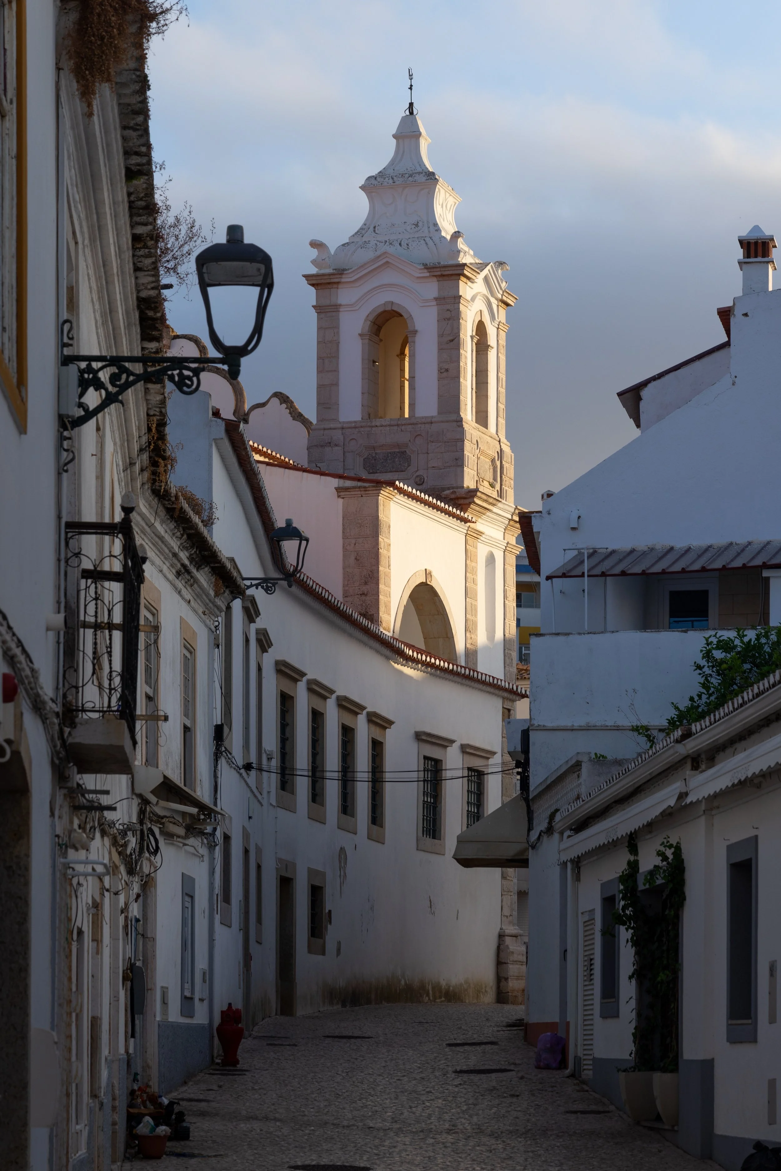 A quiet cobblestone street in a historic European town with white buildings and a church tower with a clock and spire. The sky is partly cloudy, and there are vintage-style street lamps along the street.