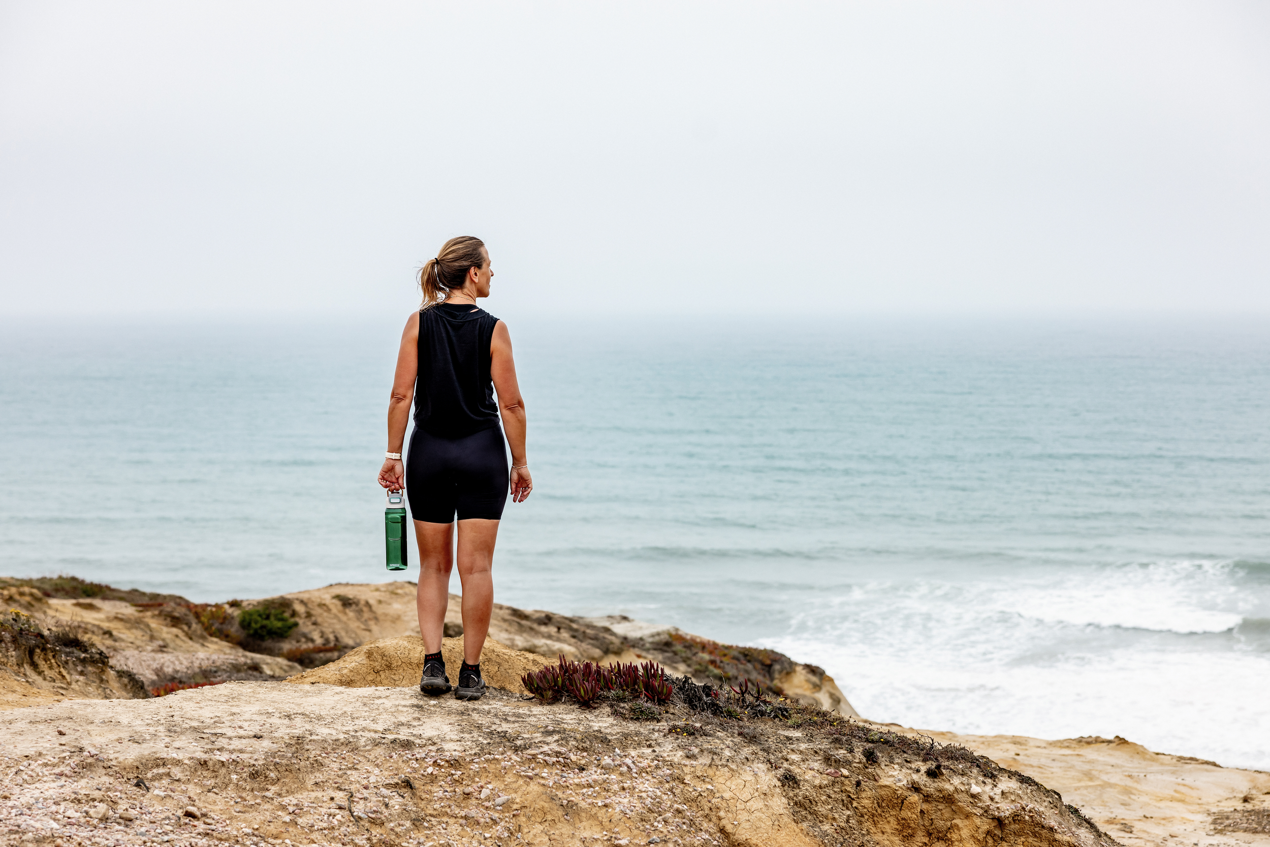 A woman dressed in athletic clothing stands on a rocky coastal landscape, looking out at the ocean with an overcast sky.