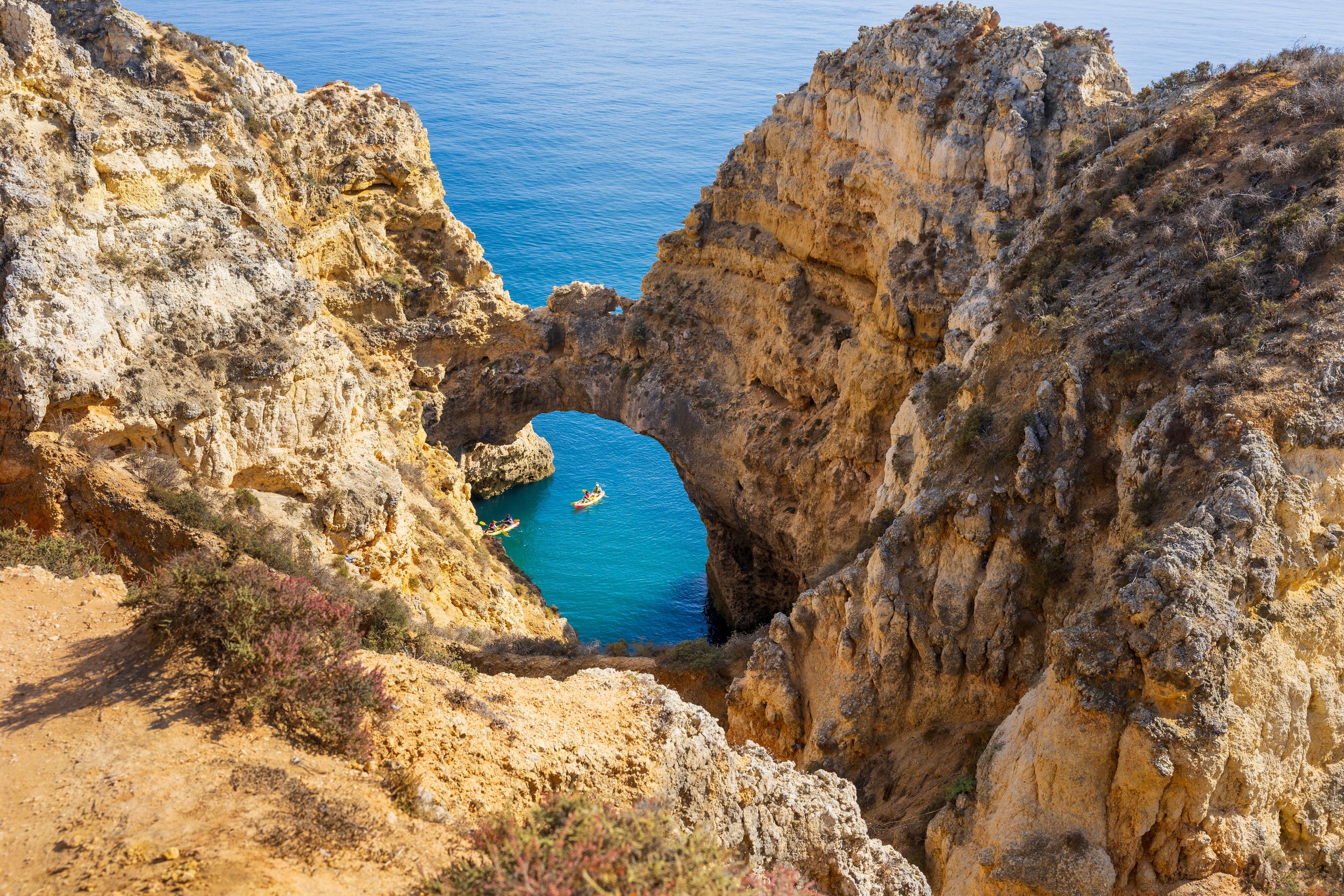 Rocky coastal cliffs with a natural arch formation over blue ocean and a small boat with people in water below.