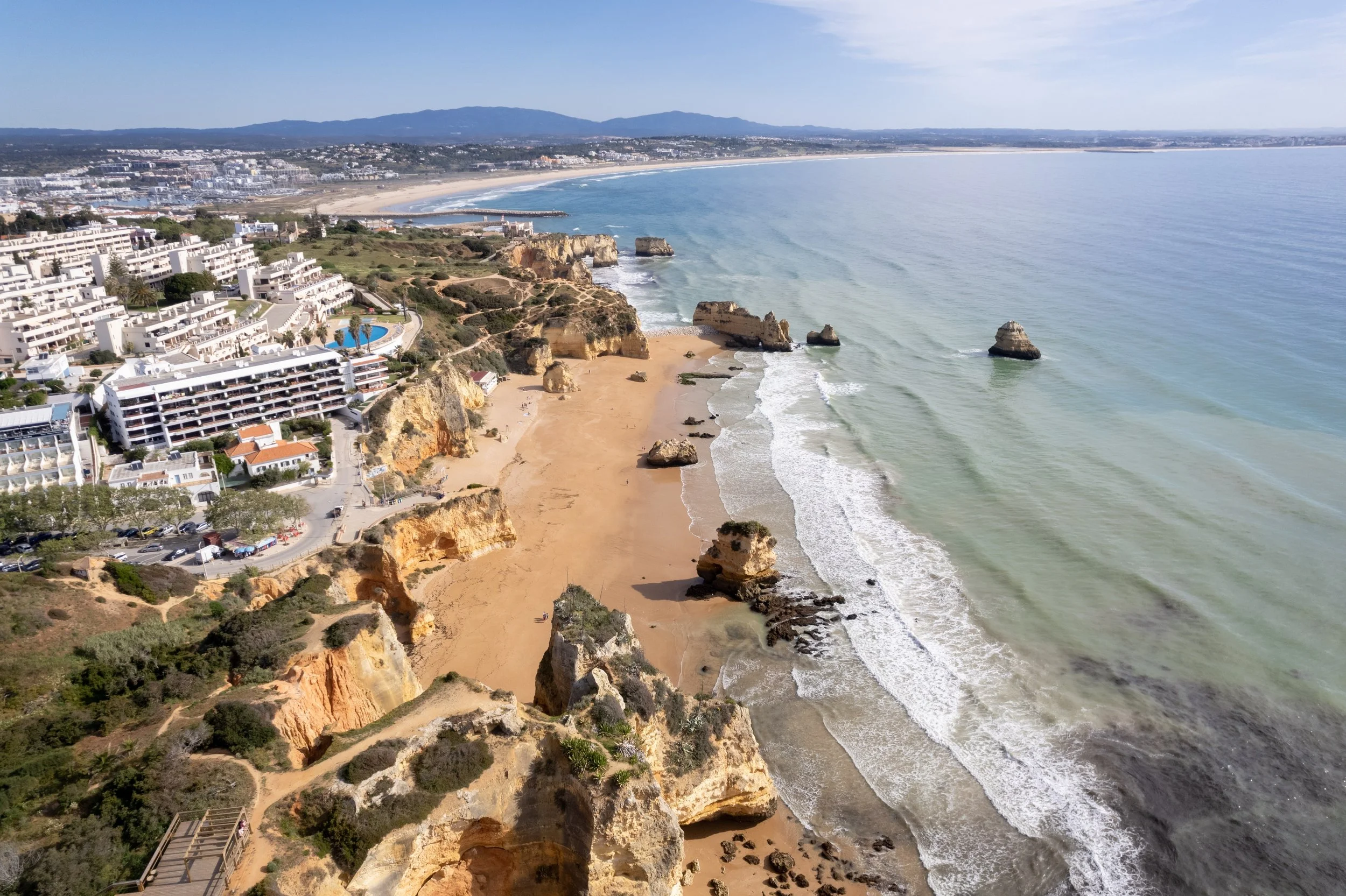 Aerial view of a coastal city with white buildings, sandy beaches, rocky cliffs, and the ocean, likely taken from a drone or high vantage point.