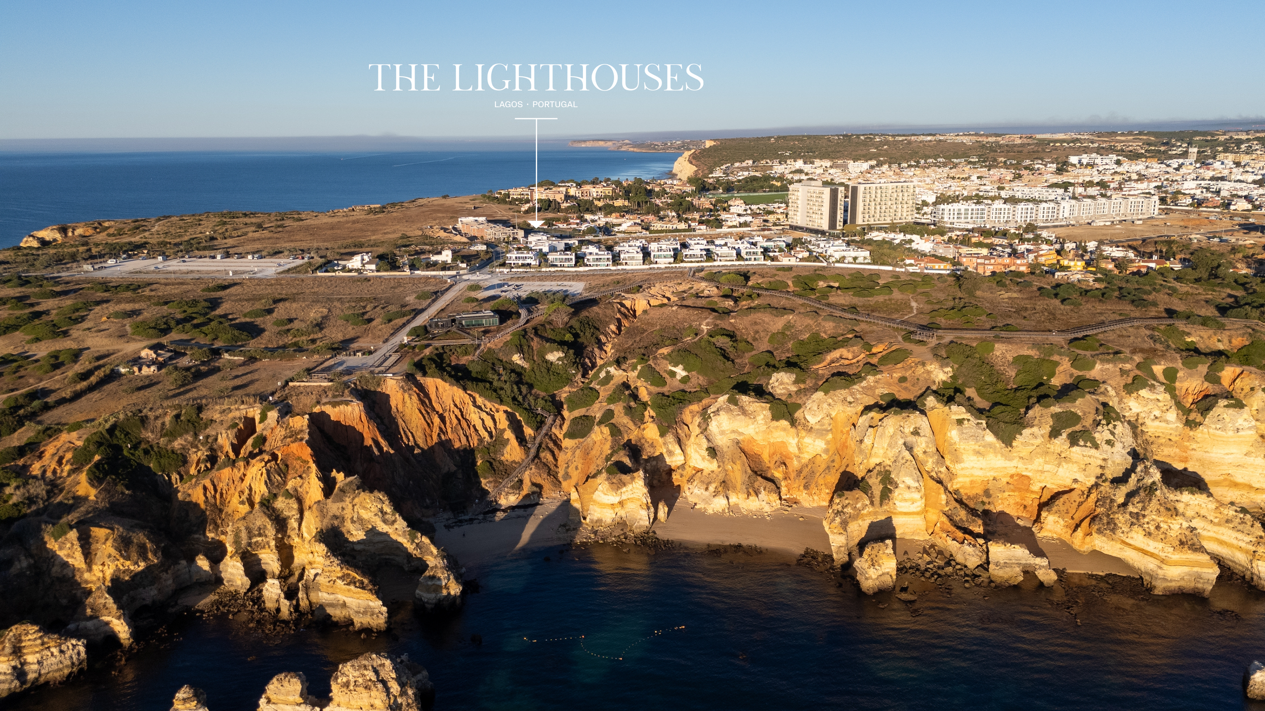 An aerial view of cliffs along the coast in Lagos, Portugal, with residential buildings and the Atlantic Ocean in the background.