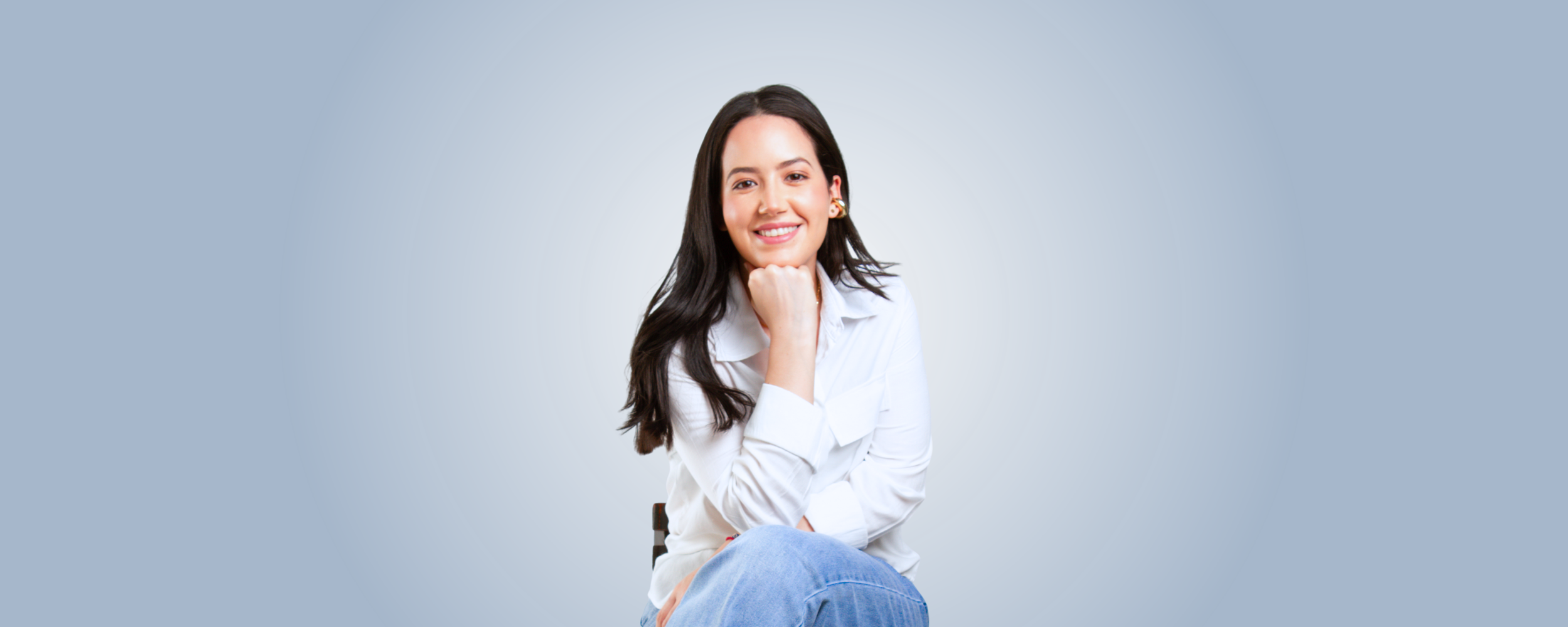 A woman with long dark hair, wearing a white shirt and blue jeans, smiling and resting her chin on her hand, sitting against a light grey background.