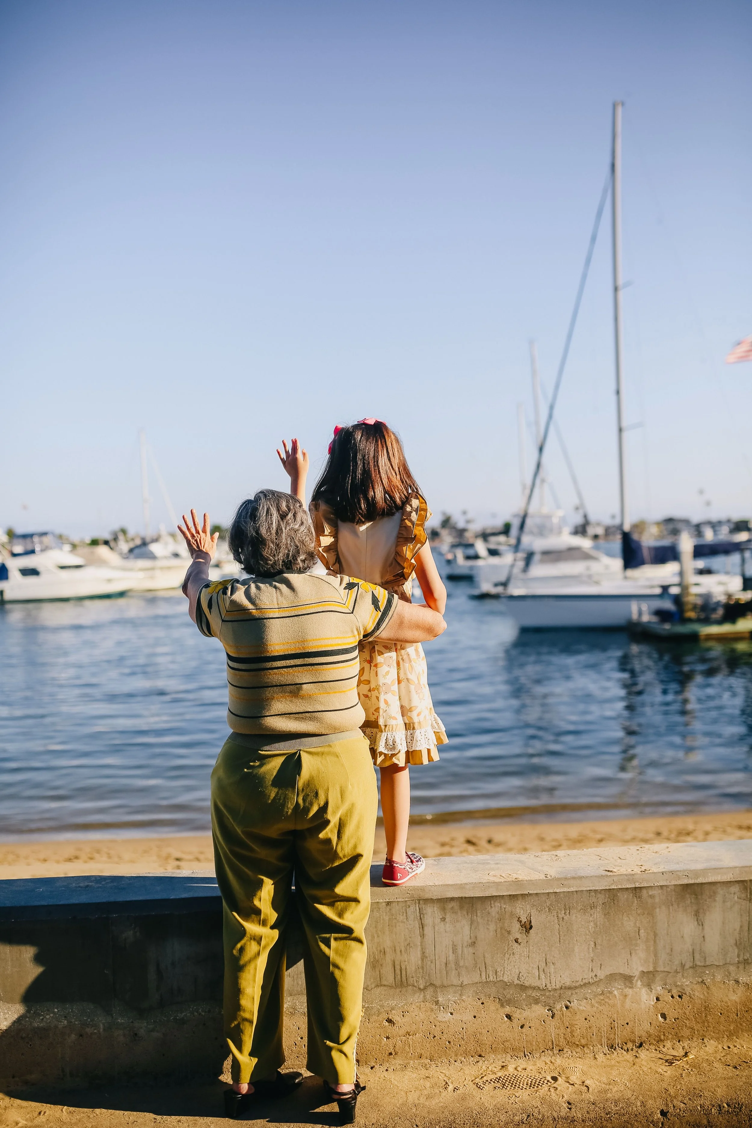 An older woman and a young girl are standing near a waterfront with boats docked in the background. The woman is holding the girl up in the air, and the girl is waving. The scene takes place in the late afternoon or early evening, with clear skies and calm water.