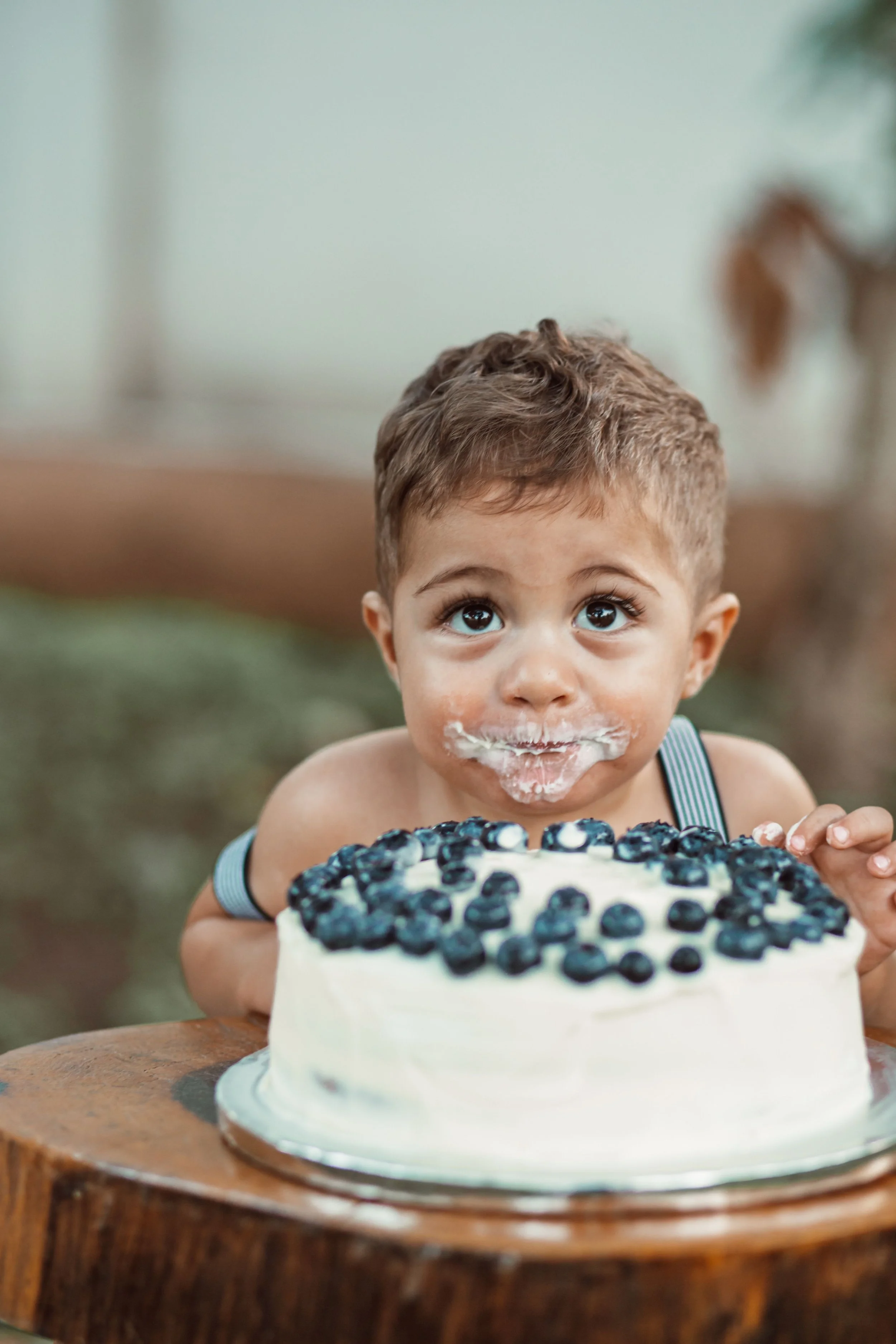 A young child with brown curly hair, big expressive eyes, and frosting on his face is excitedly looking at a blueberry cake in front of him during his first birthday celebration.