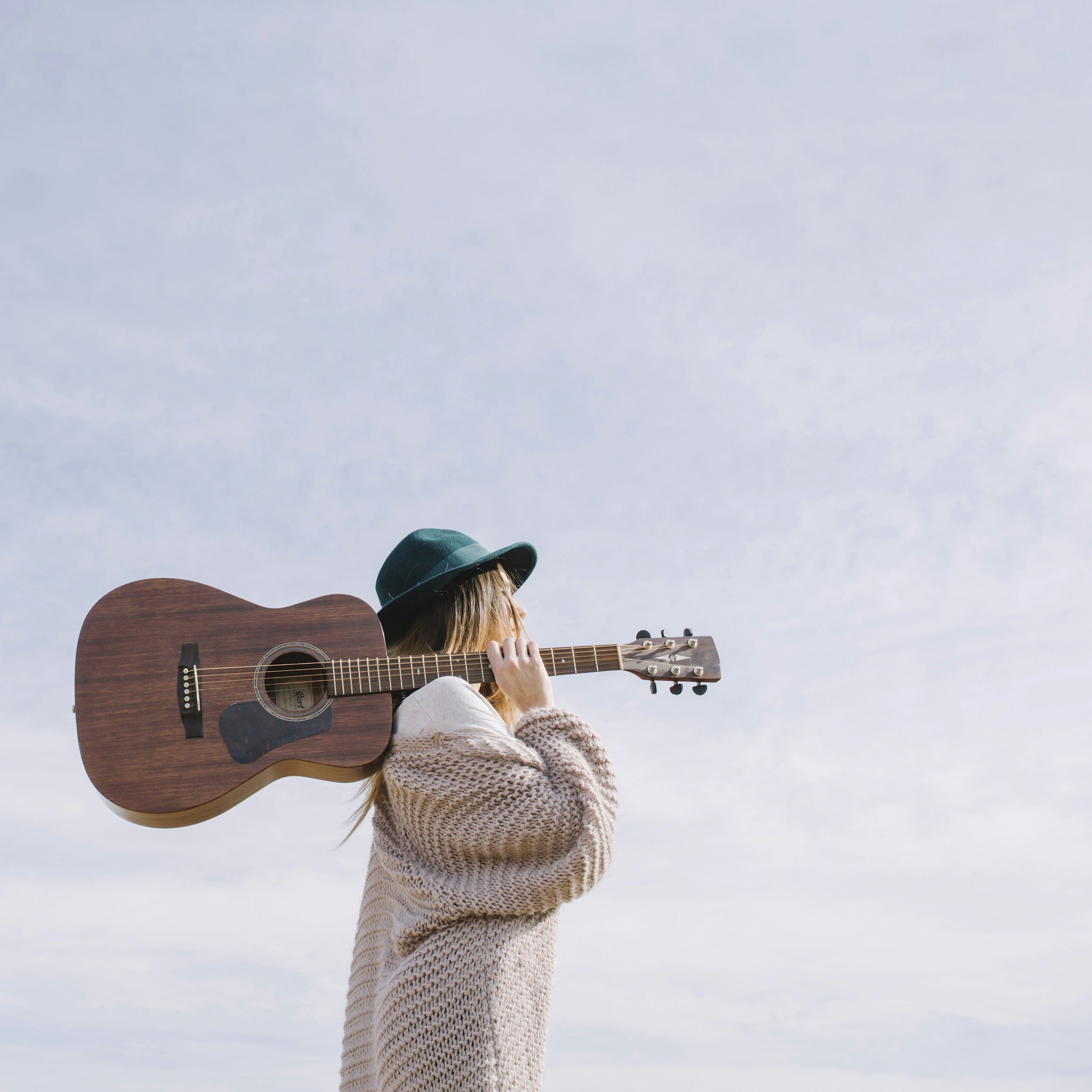 A person wearing a beige sweater and a teal hat, holding a wooden acoustic guitar, facing away against a cloudy sky.