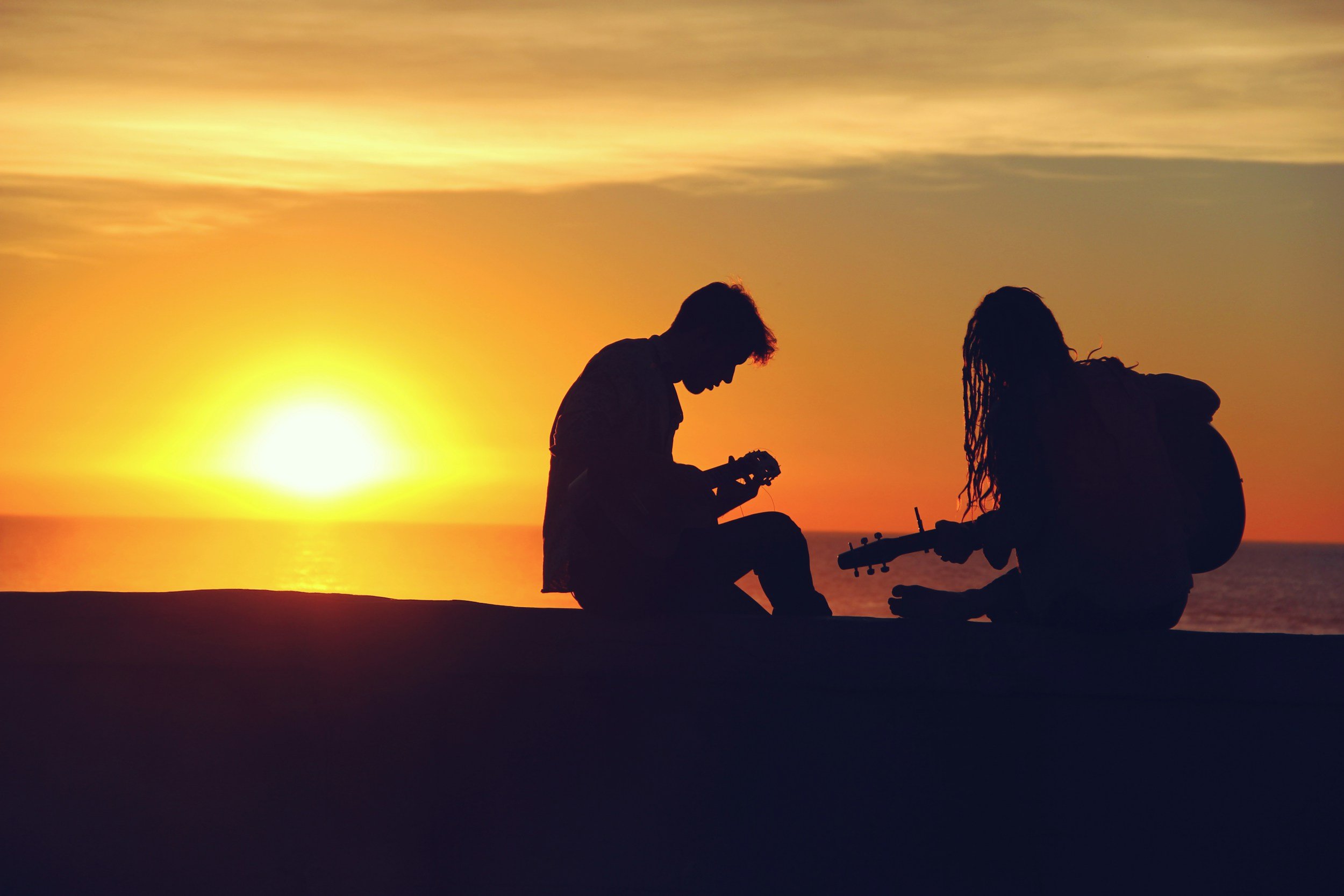 Two people sitting by the water at sunset, one playing guitar and the other holding a small object, with the sun setting on the horizon and a partly cloudy sky.