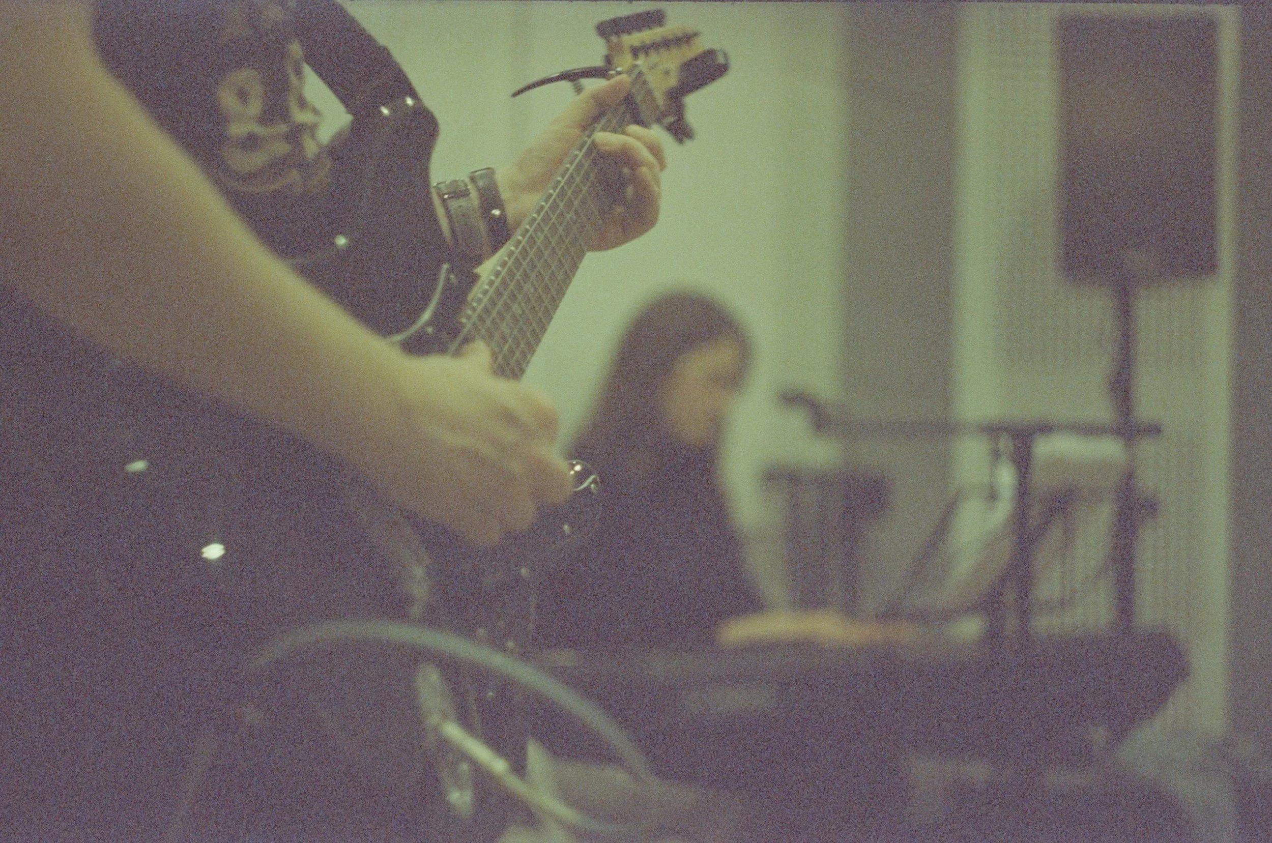 A person playing an electric guitar in a room, with musical equipment visible in the background.