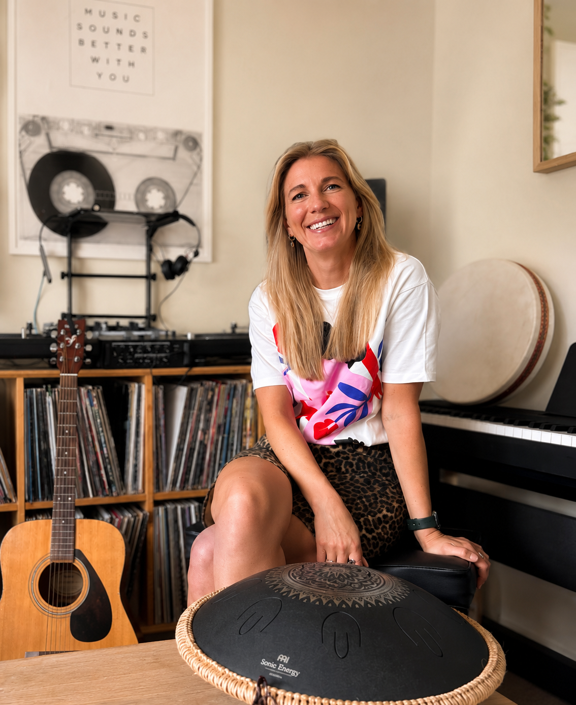 A woman with long blond hair smiling, sitting in a music studio, surrounded by musical instruments and records, with a guitar and handpan in front of her.