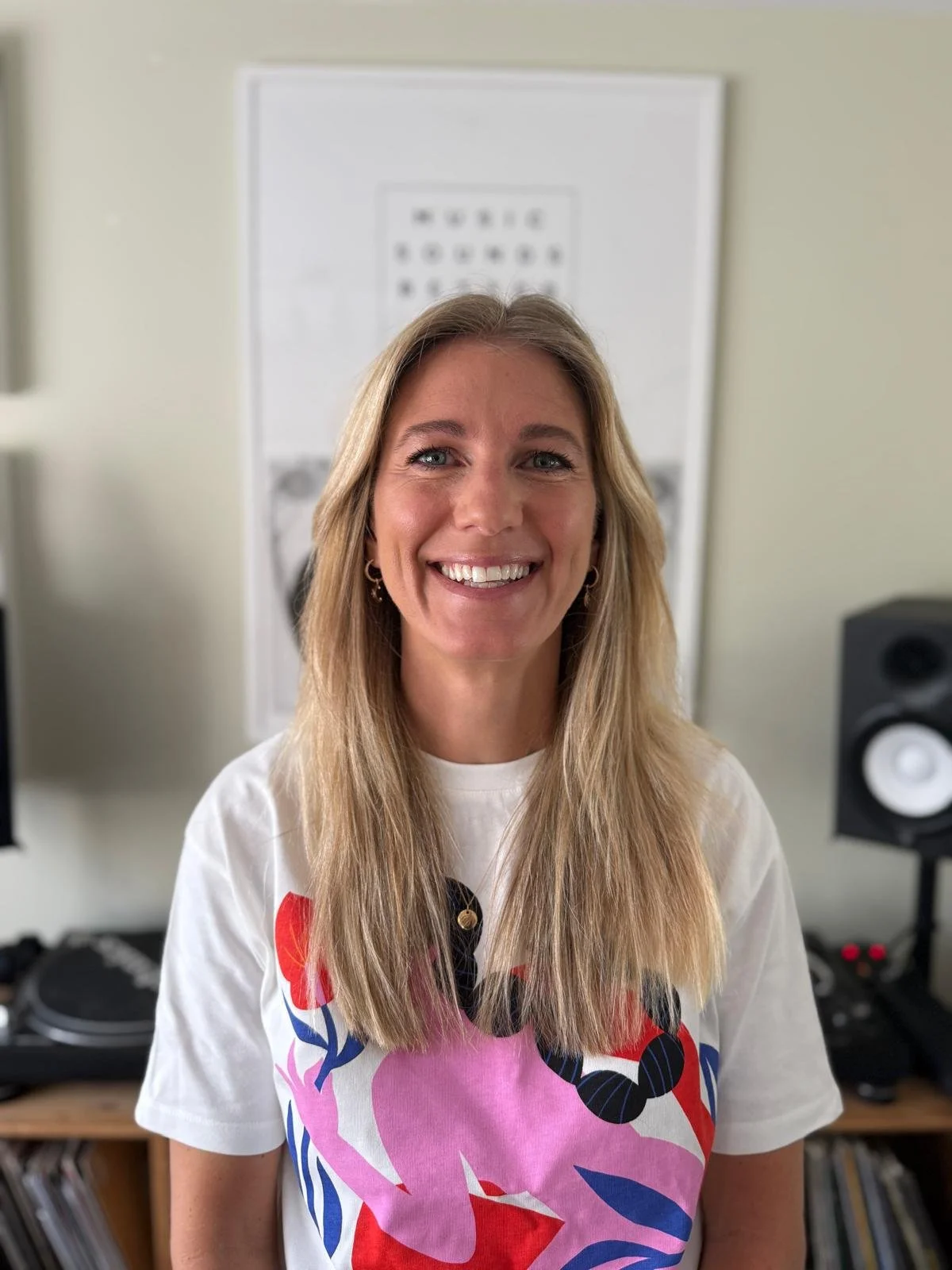 Smiling woman with long blonde hair standing in a room with audio equipment in the background.