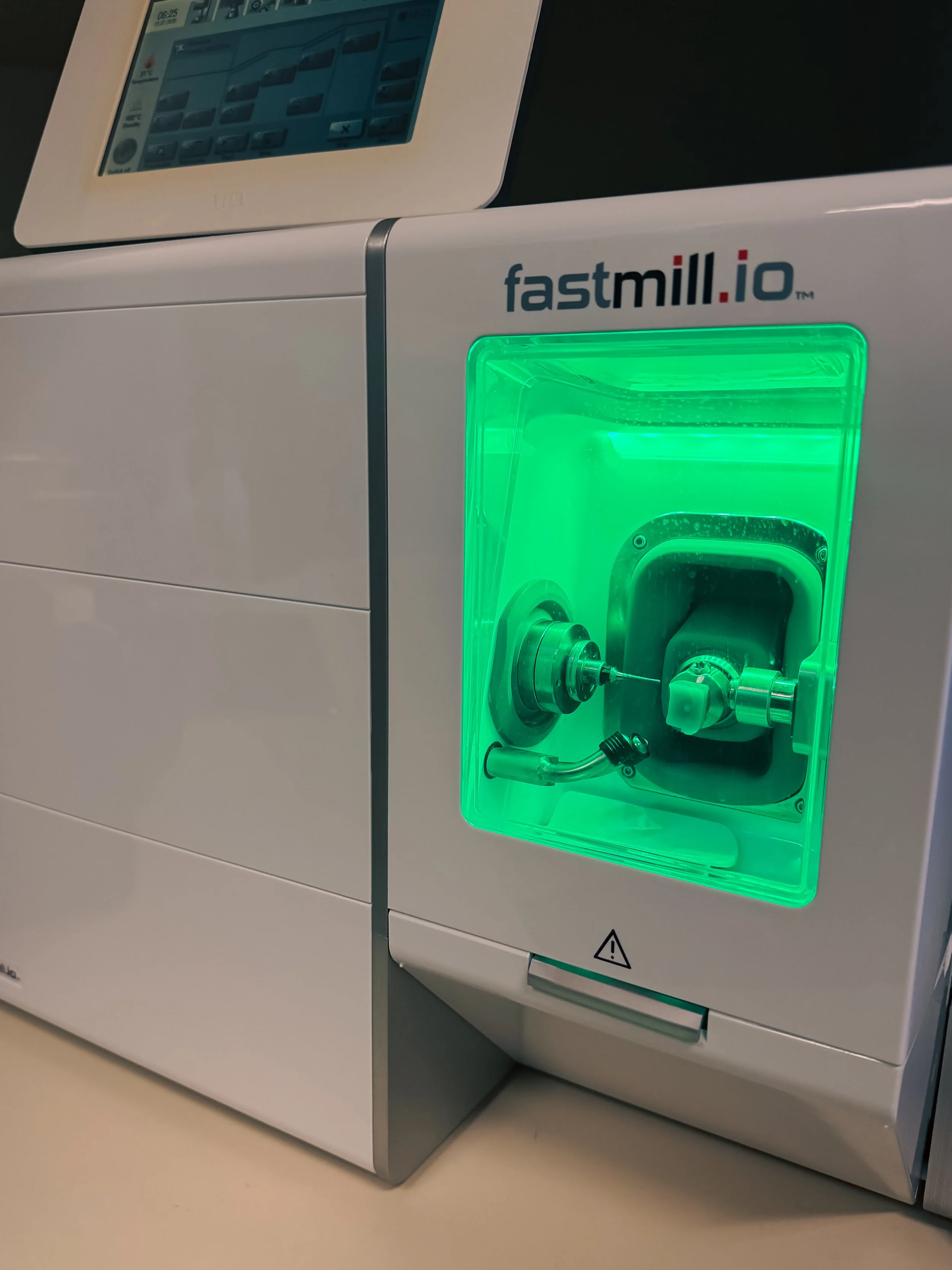 Close-up of a white machine labeled 'fastmill.io' with a green-lit access port containing metal parts and some black tubing, located in a clinical or laboratory setting.