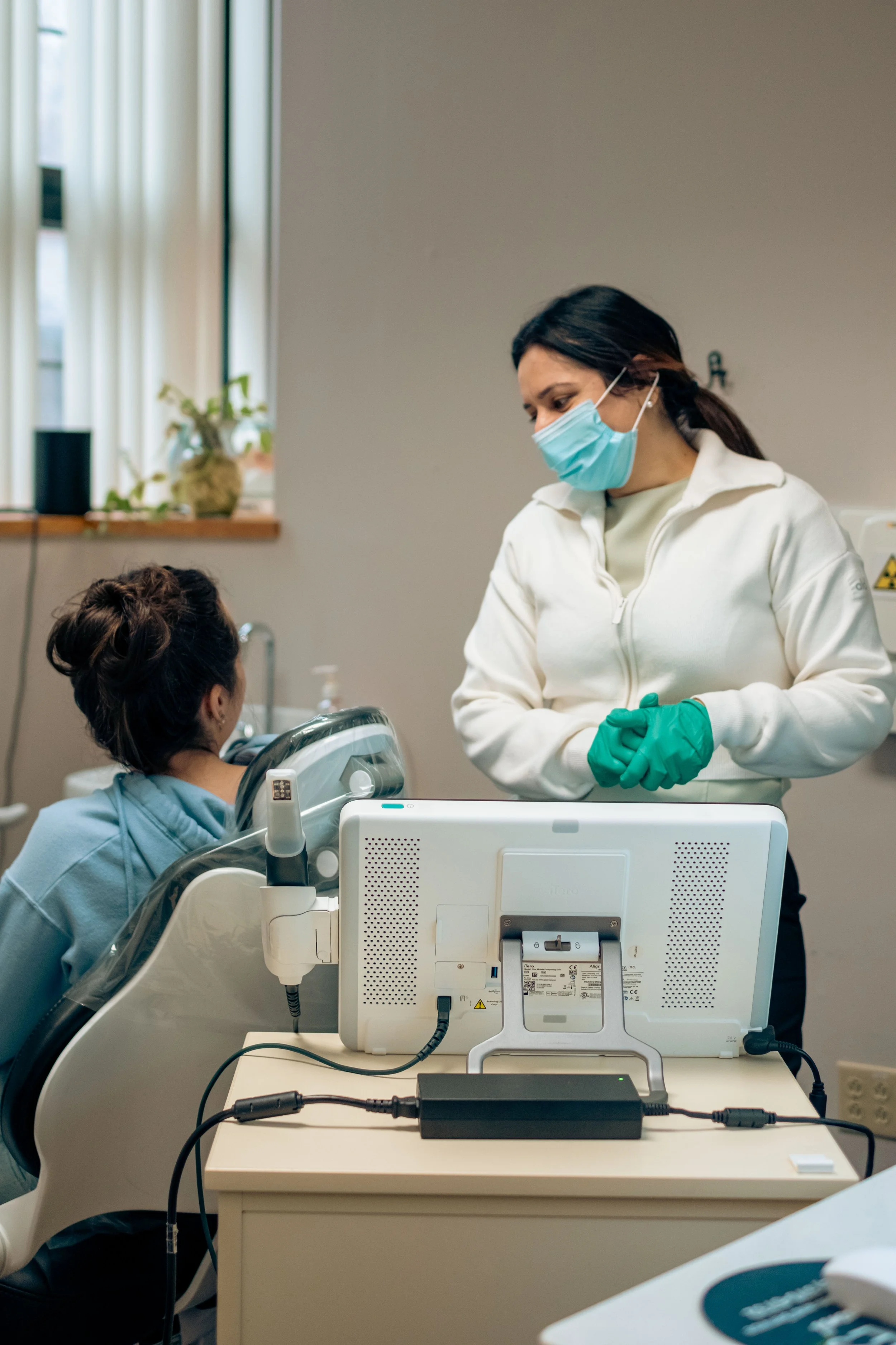 Medical professional wearing a face mask and gloves converses with a patient in a hospital room. The patient, seated in a hospital bed, has a hair bun and wears a hospital gown. Medical equipment, including a monitor, is visible in the foreground.