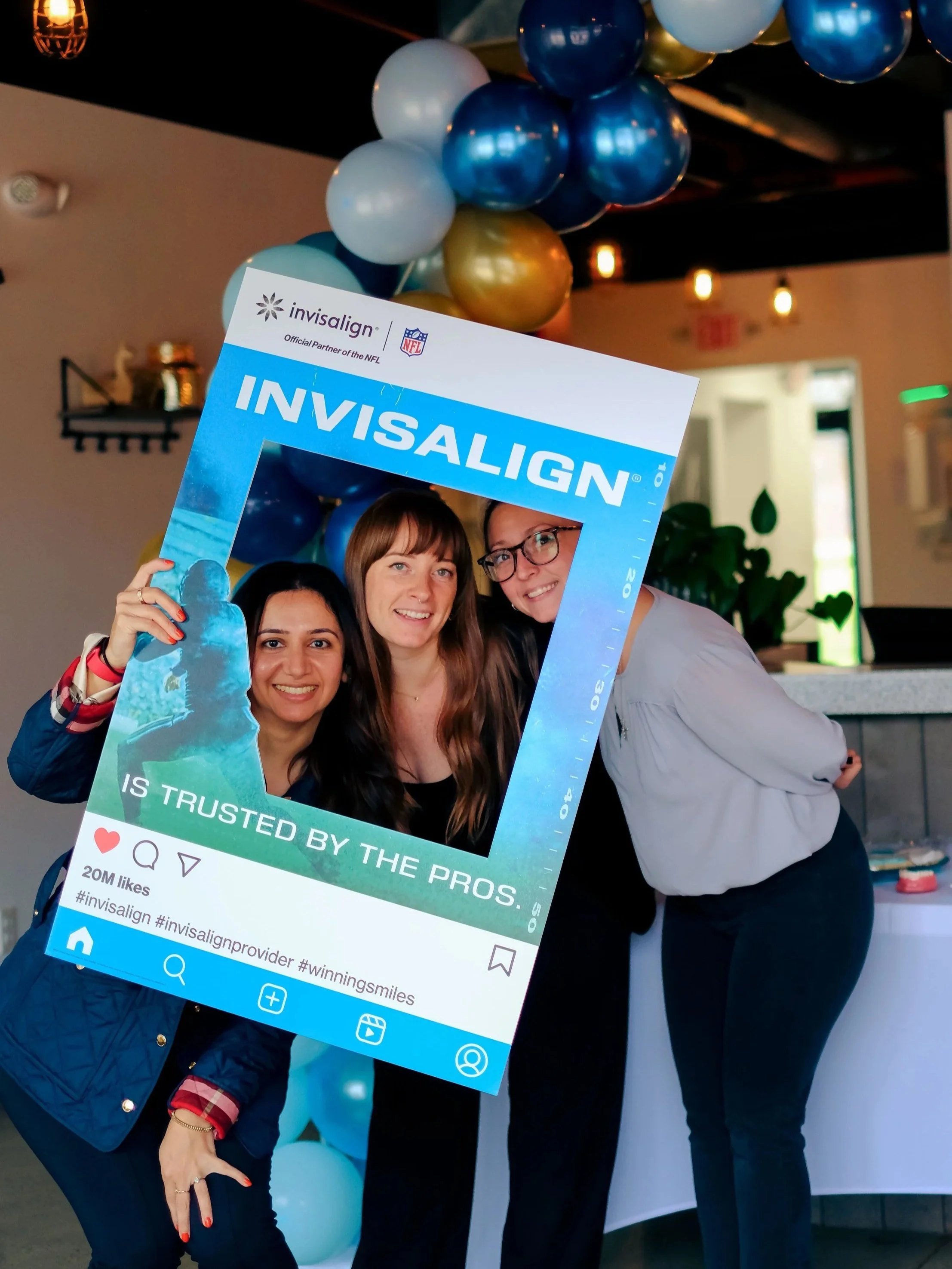 Three women and one man holding a large fake social media post frame at an indoor event, with blue and gold balloons overhead. The frame has Facebook style icons and the word 'Invisalign' at the top.