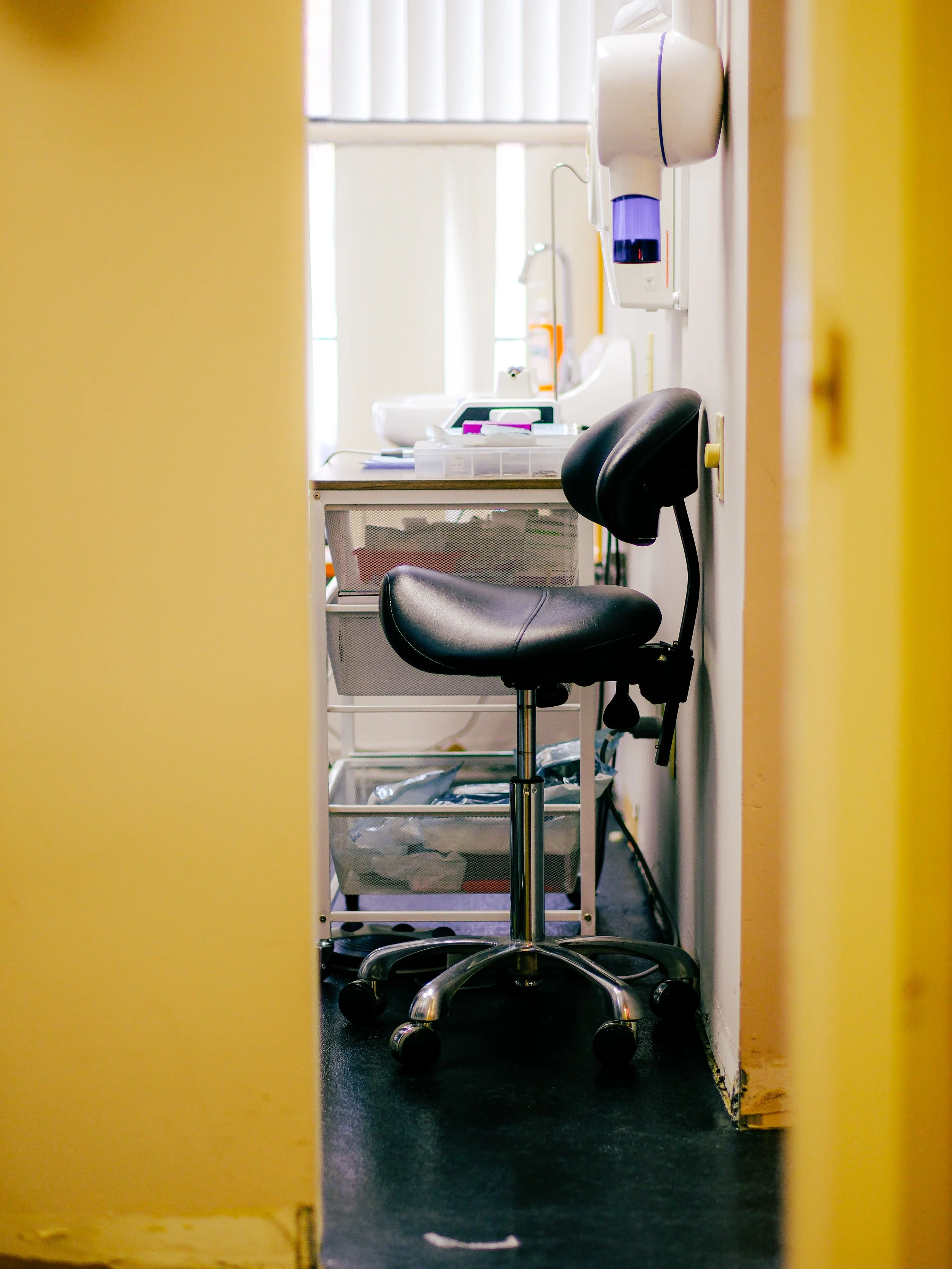 View through a partially opened yellow door showing a medical examination room with a black office chair, medical equipment, and supplies on a cart.