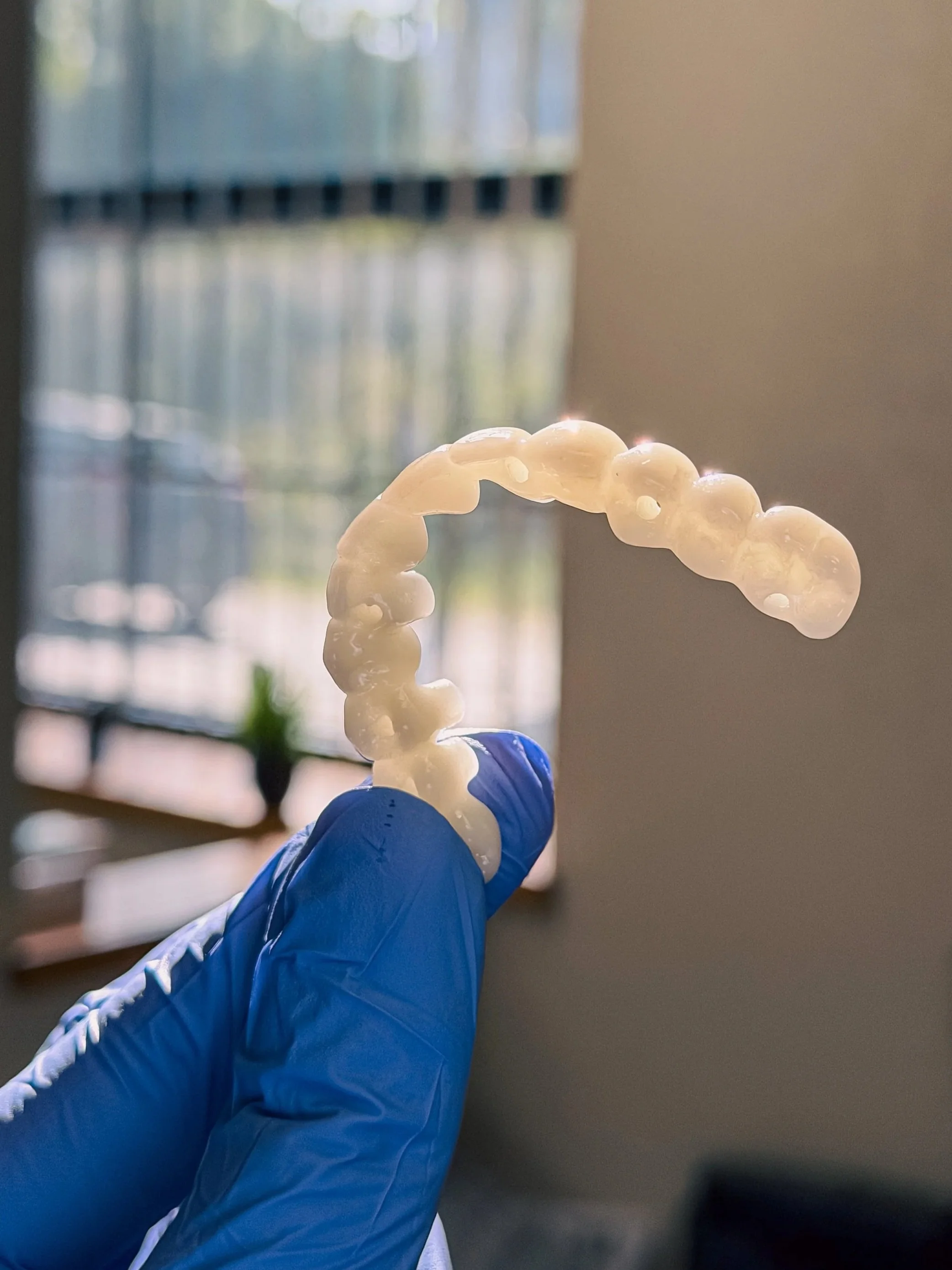 Close-up of a blue-gloved hand holding a clear dental aligner or retainer near a window with blinds, sunlight reflecting off the plastic.