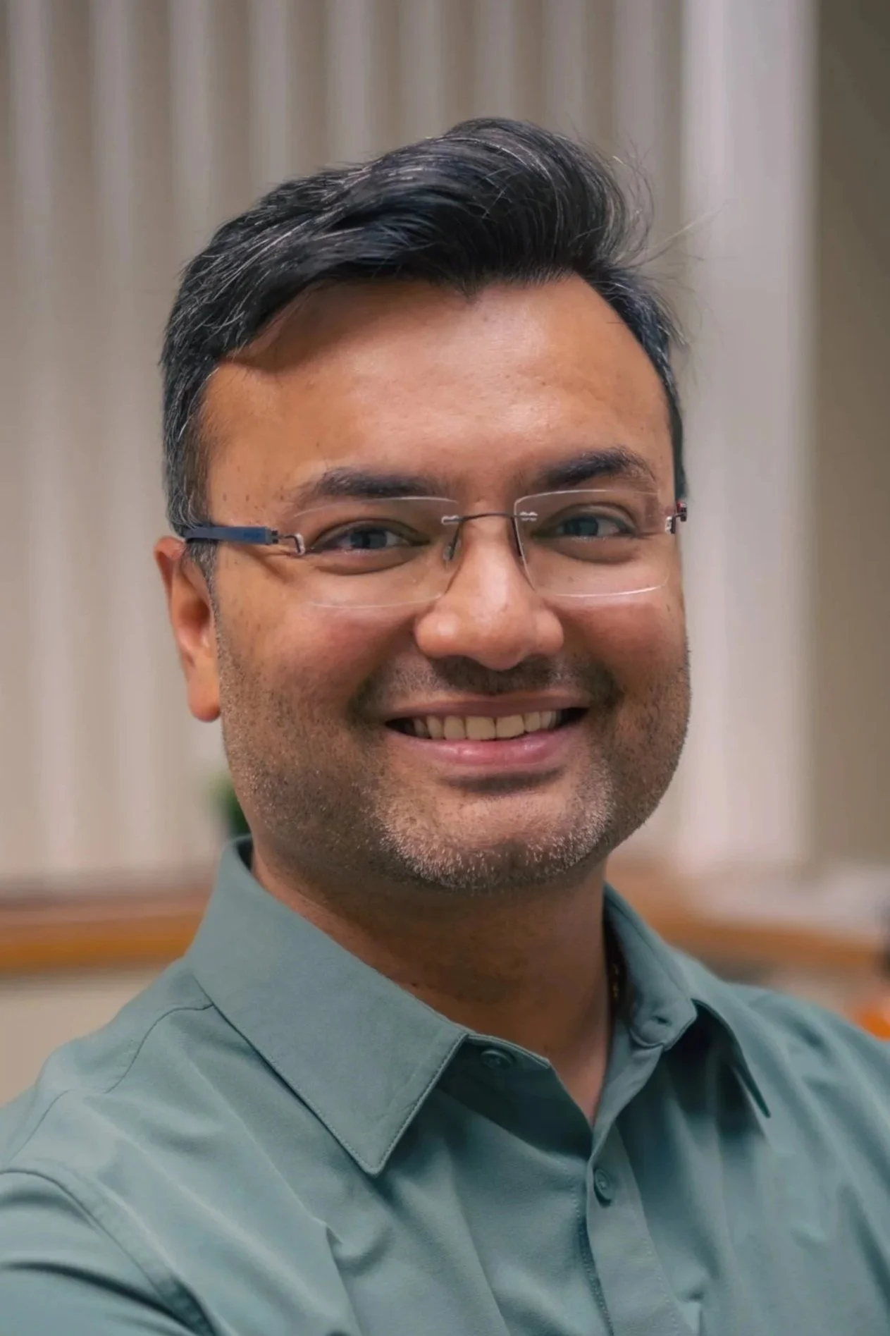 A smiling man with glasses, dark hair with some gray, wearing a green collared shirt, in a softly lit room with cream curtains in the background.