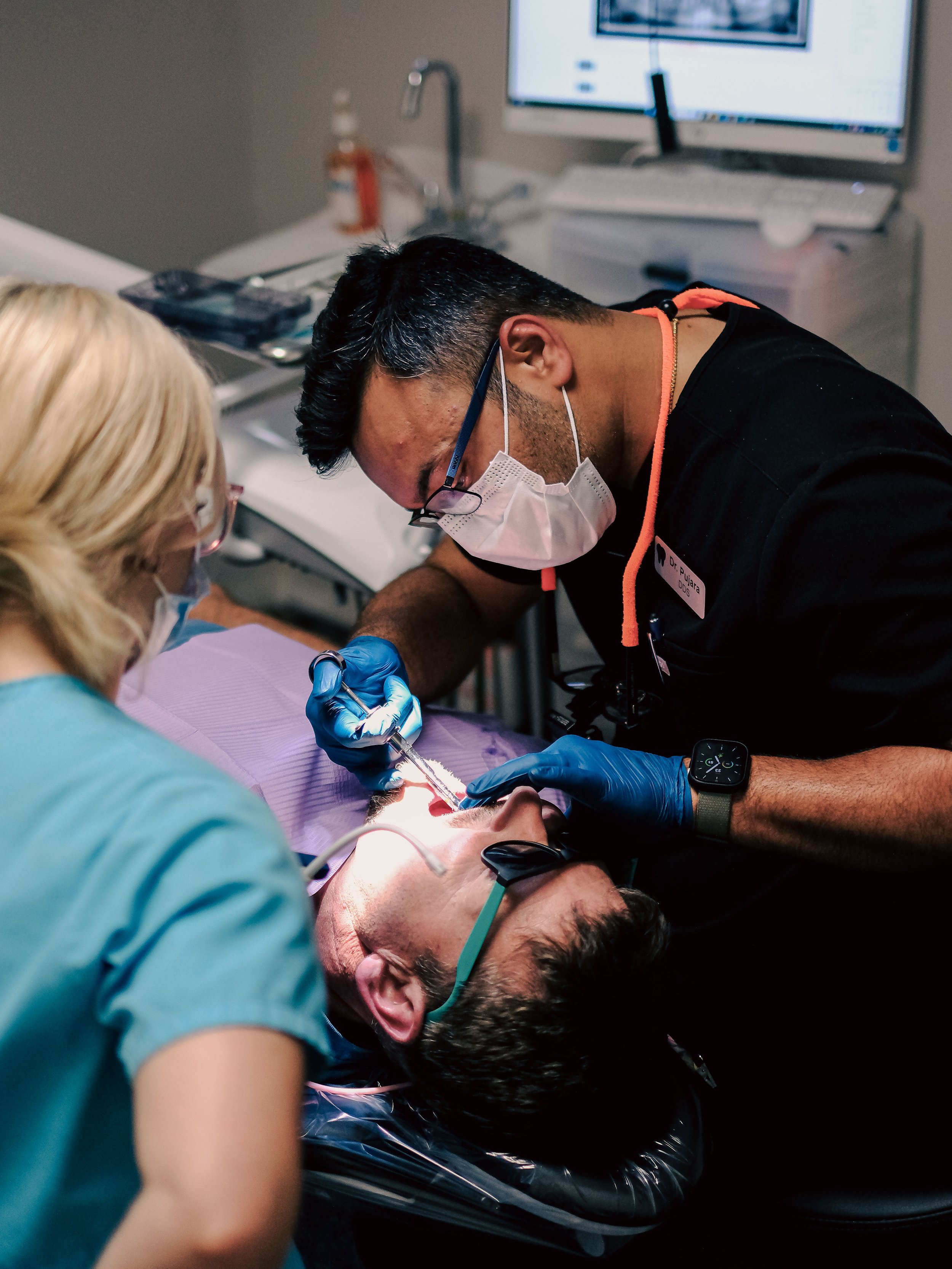 Dentist performing a dental procedure on a patient lying in a dental chair with another dental professional assisting.