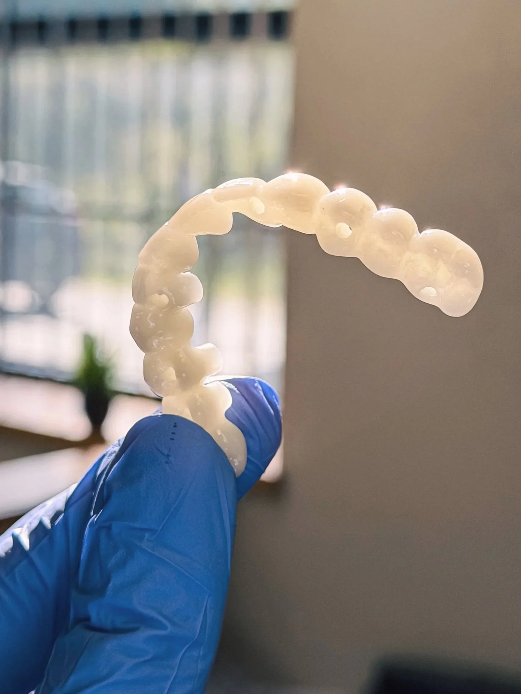 A gloved hand holding a clear aligner or dental retainer in front of a window with blurry vertical blinds in the background.