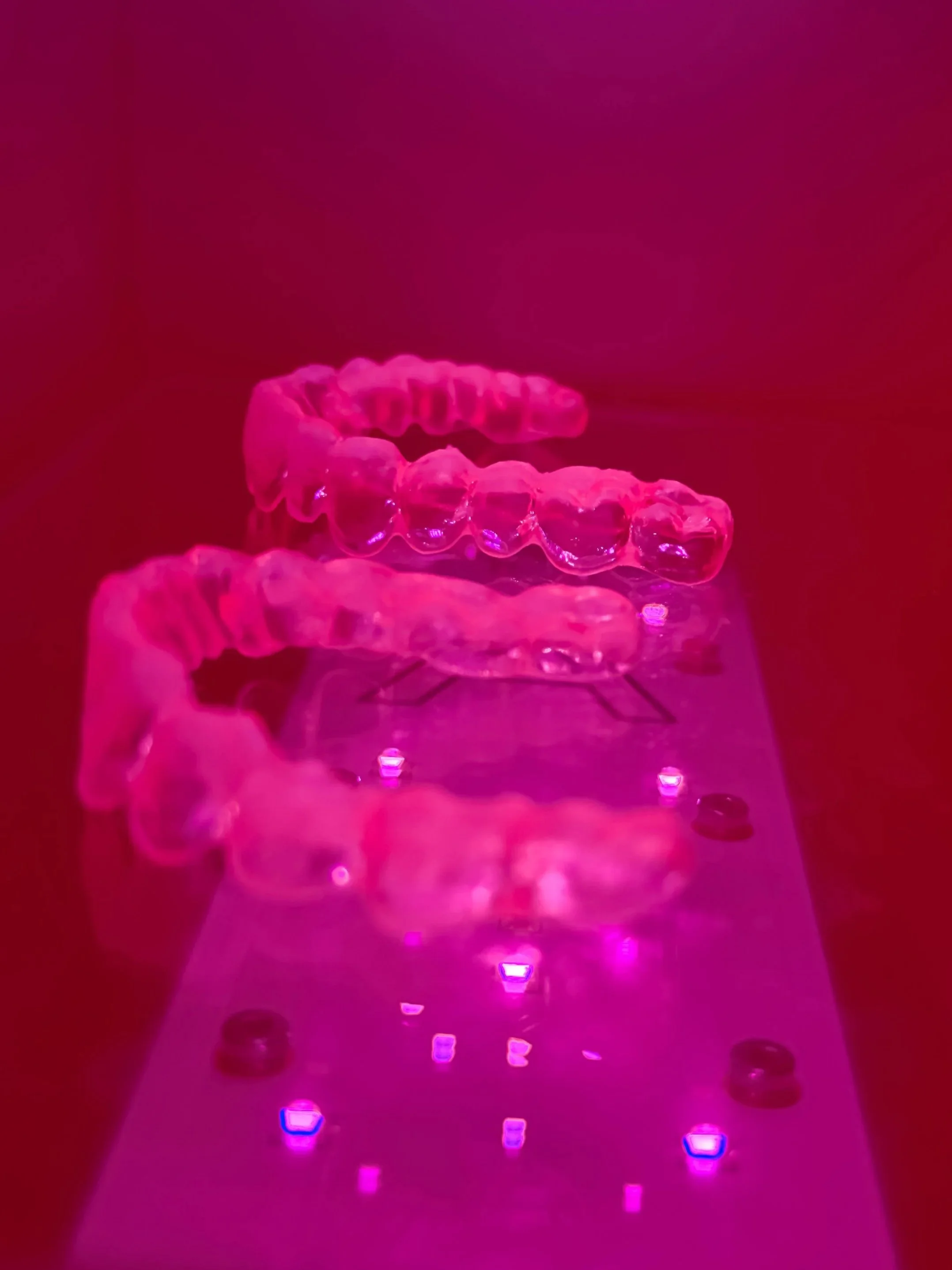 Close-up of a clear aligner dental tray illuminated with pink and purple lighting on a reflective surface.