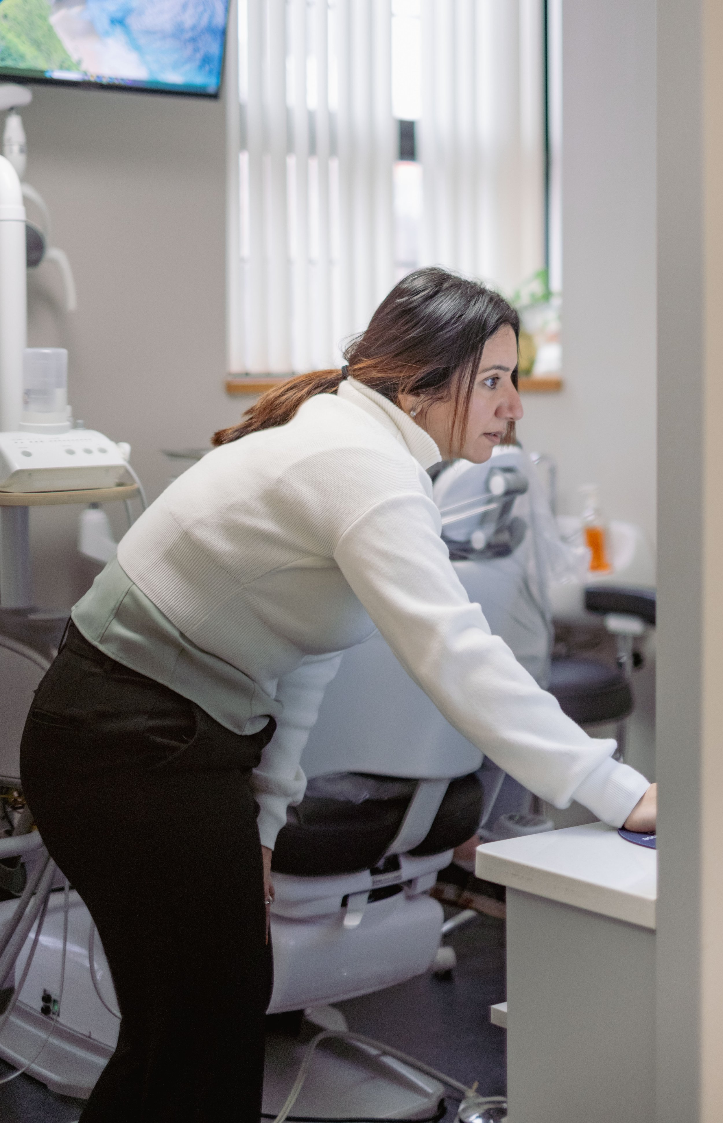 A woman leaning over and working at a desk in a medical or clinical setting, with medical equipment and a monitor in the background.