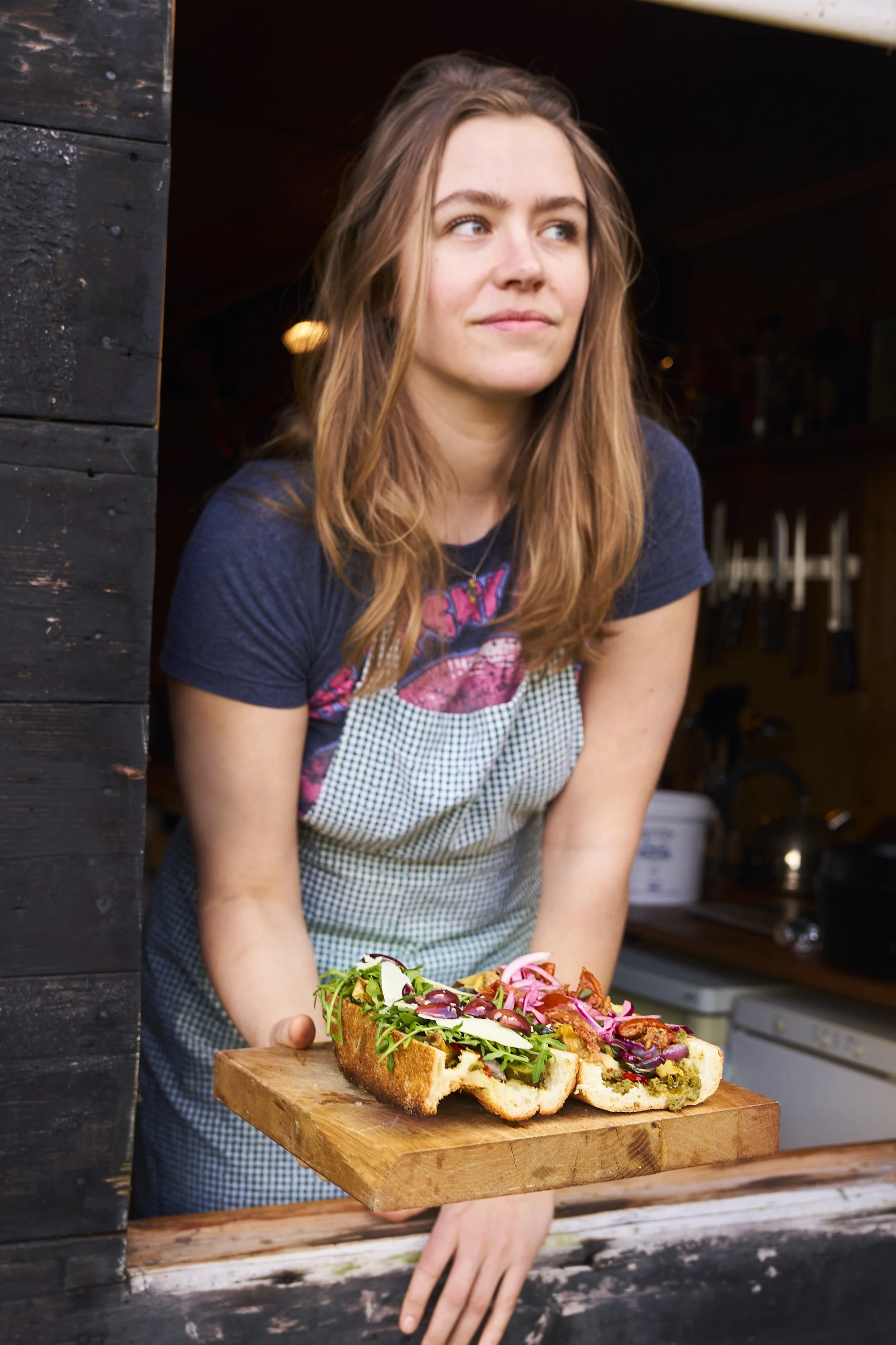 Een vrouw met bruin haar in een blauwe T-shirt en een grof geruit schort houdt een houten plank met een grote pizza boven een houten rand.