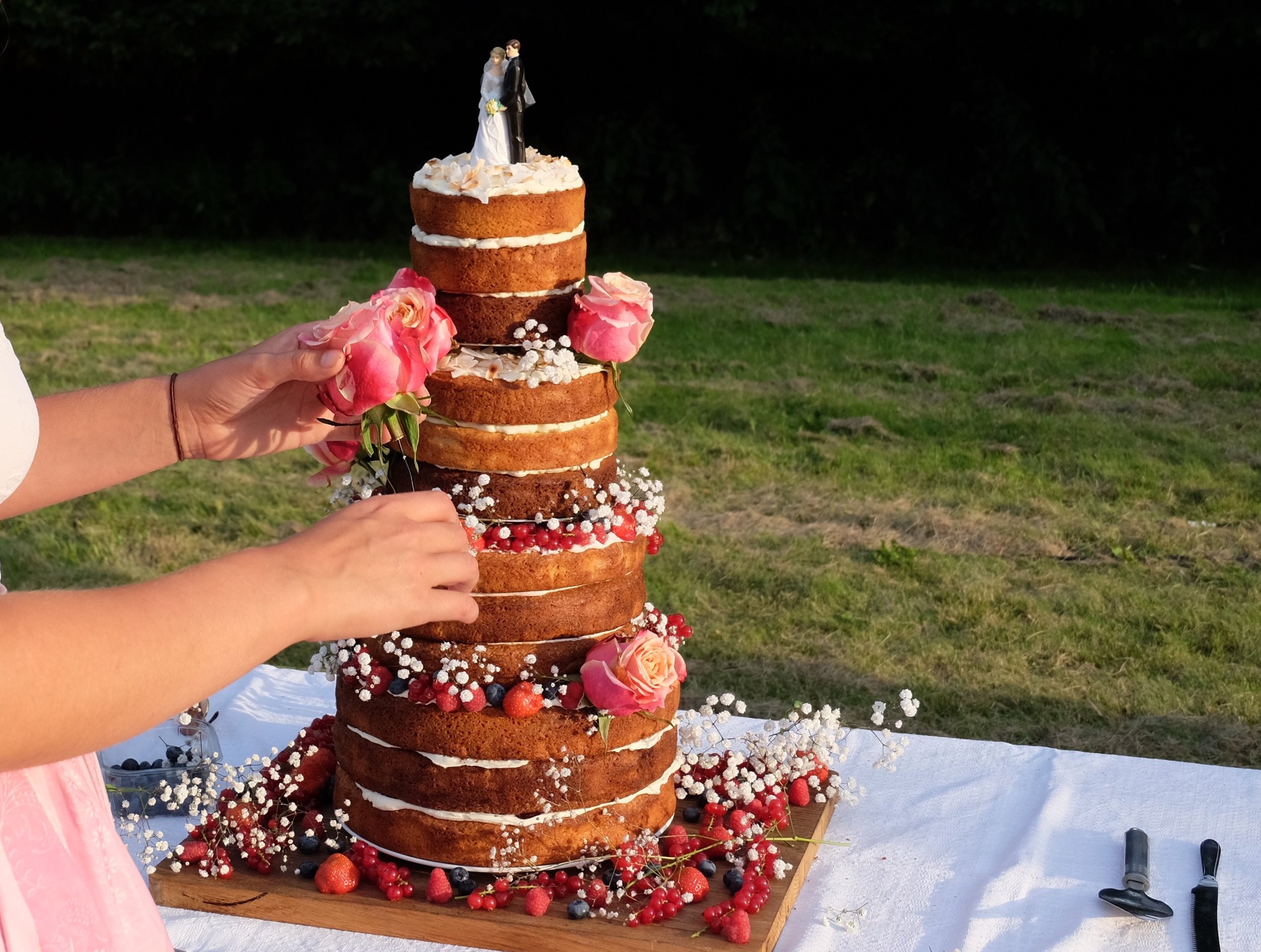Een laagjes bruidstaart versierd met bloemen en fruit, met een trouwelijkscake-topper bovenop, buiten op een wit tafelkleed.