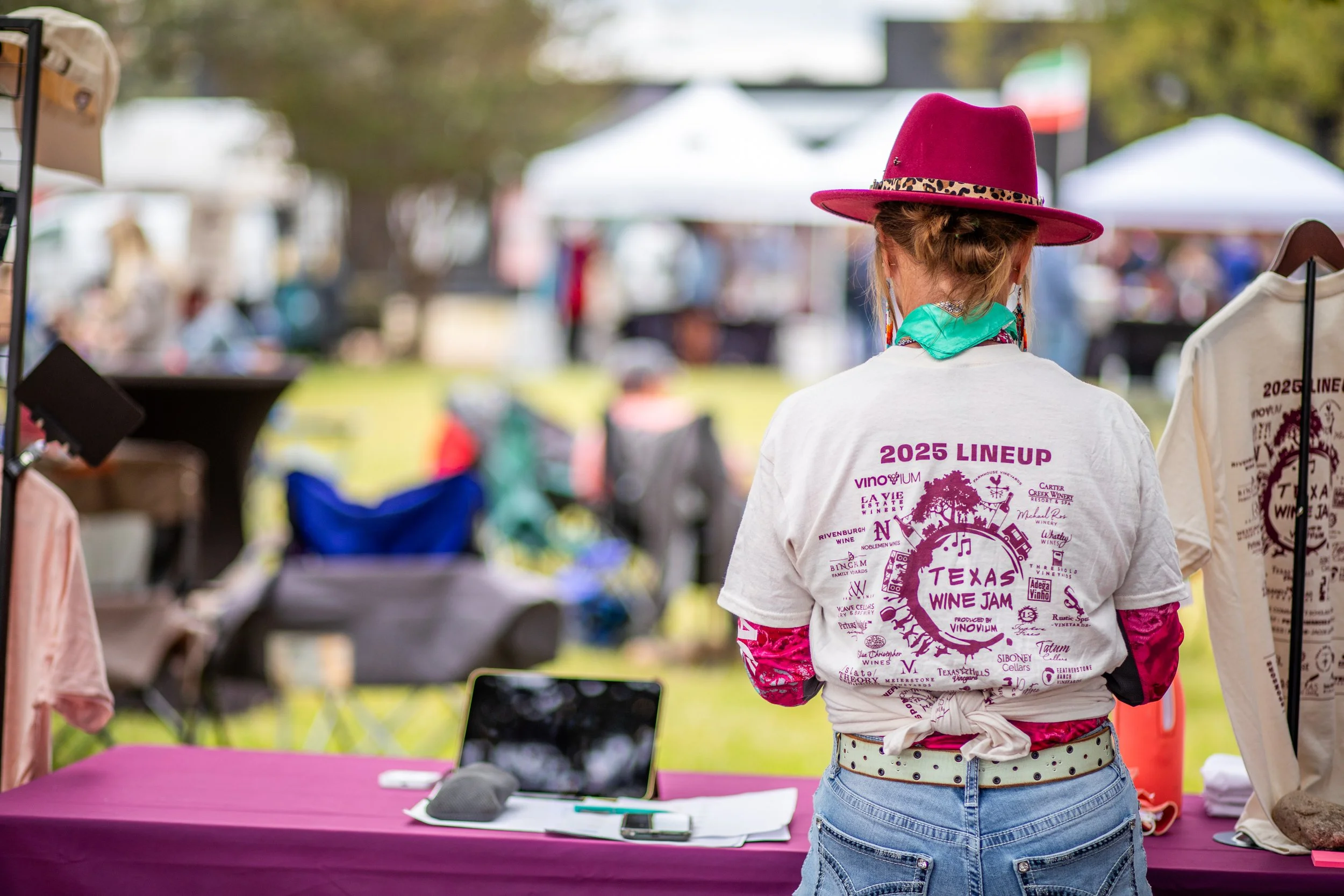 A woman wearing a pink hat with a leopard print band, a white T-shirt with 'Texas Wine Jam' and various winery logos, and jeans standing at an outdoor booth during a festival or event, with blurred tents and people in the background.