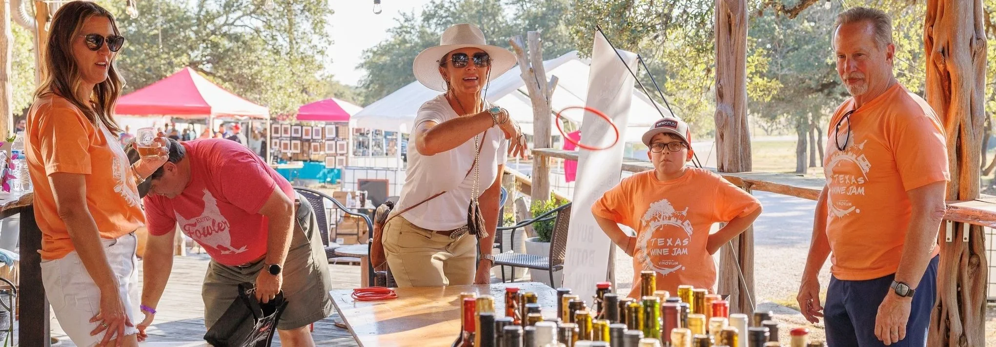 A group of five people, including three adults and one child, at an outdoor event with tents. They are participating in a game involving tossing a hoop over a string while standing around a table with cans. The scene is sunny, with trees in the background, and everyone is wearing casual clothes, some with orange t-shirts.