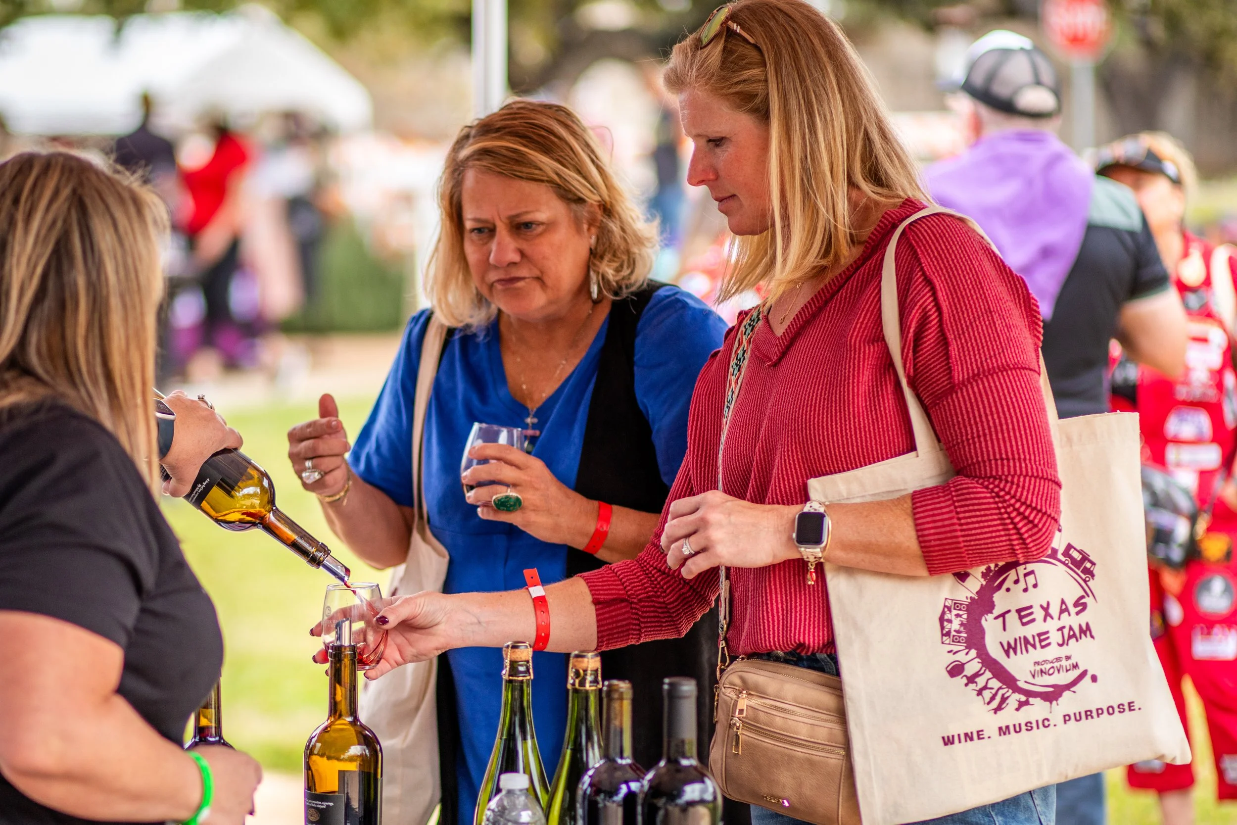 Two women are attending a wine tasting event outdoors, inspecting and pouring wine from bottles. They are engaged in conversation and appear to be enjoying the event.