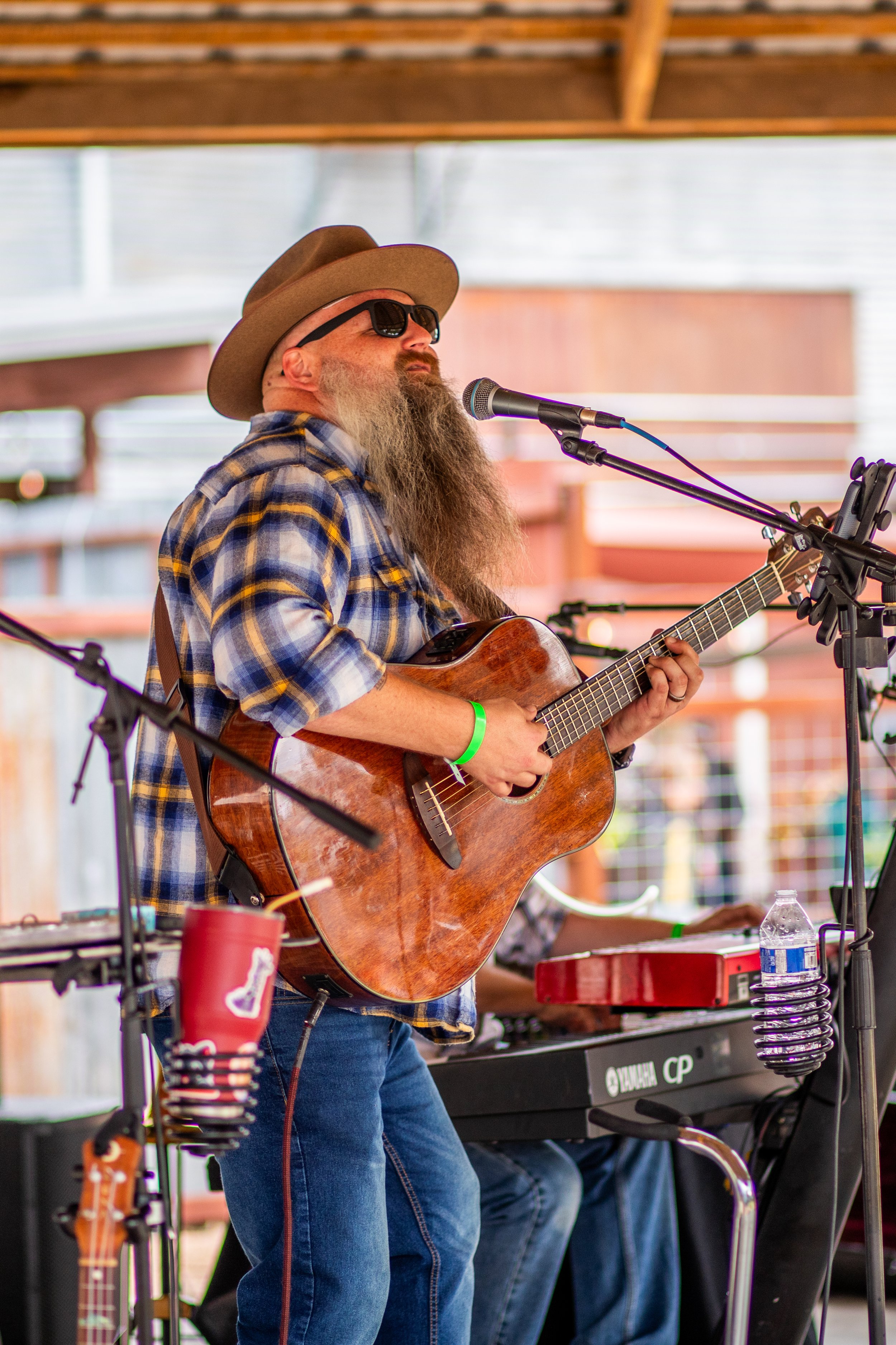 A man wearing sunglasses, a brown hat, and a plaid shirt plays an acoustic guitar during a live music performance. He stands in front of a microphone and appears to be singing or speaking. There is musical equipment and a water bottle on a table nearby.