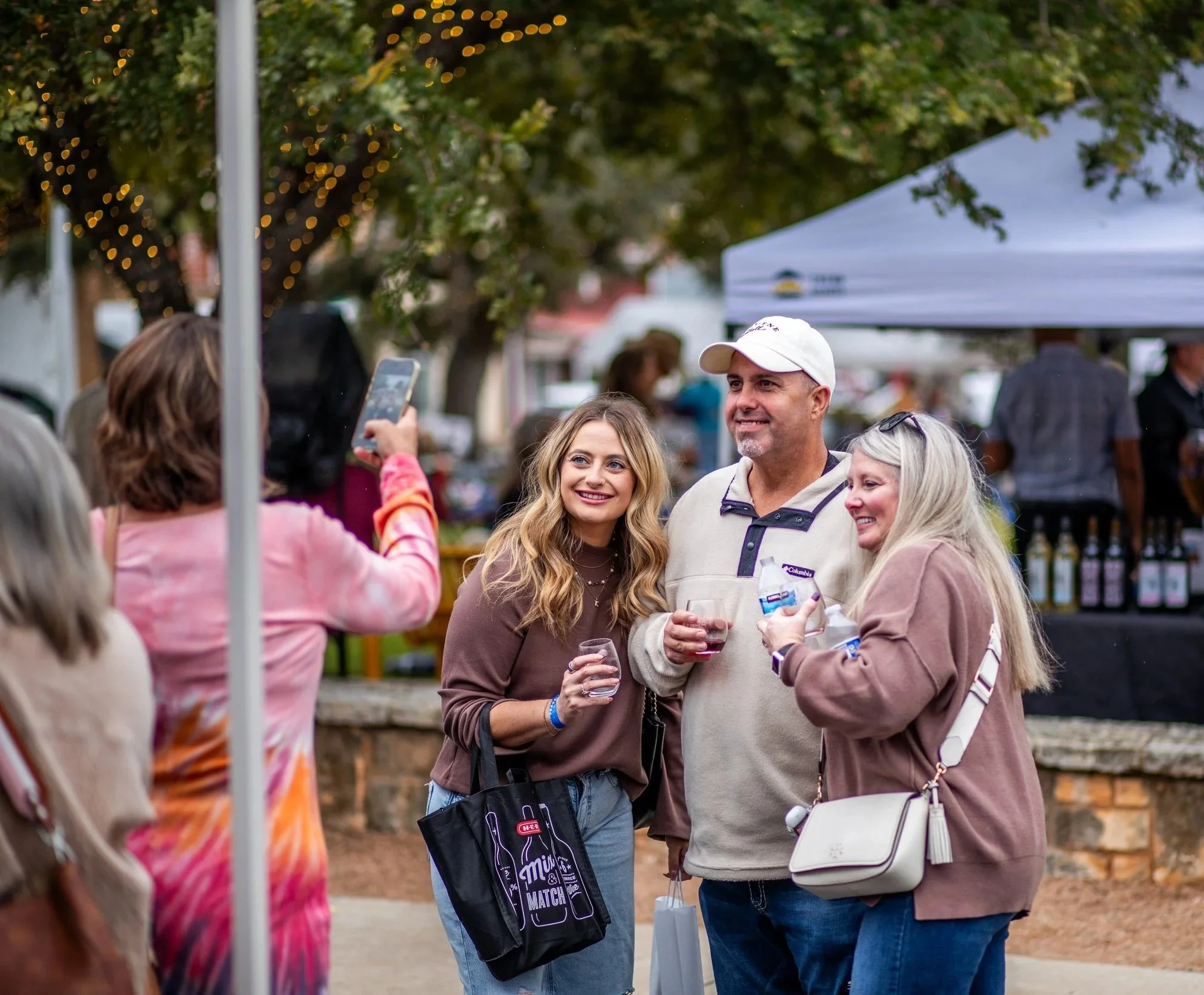 People at an outdoor event, with some taking photos and engaging in conversation, under decor with string lights and Trees in the background.