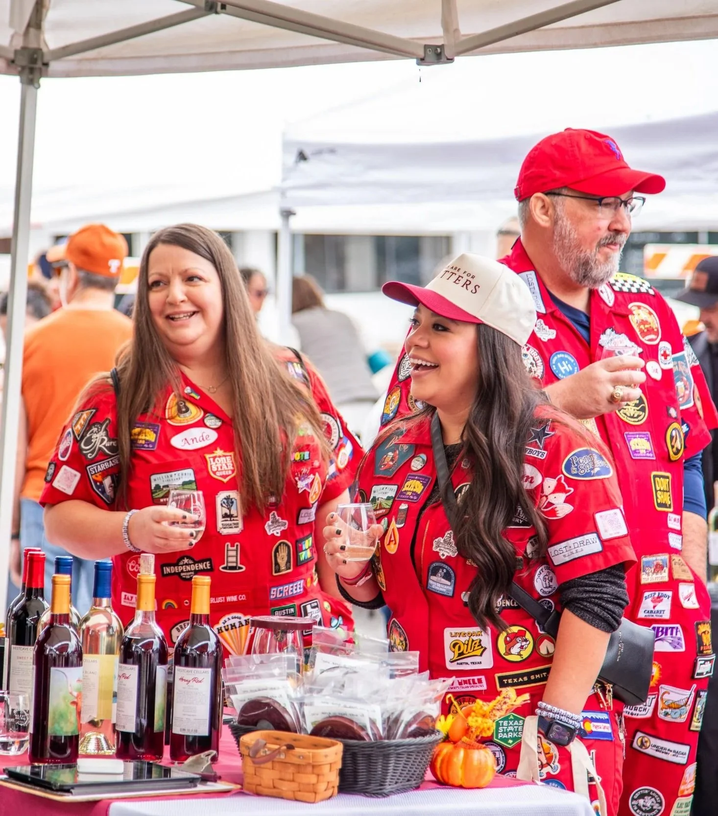 Three people at an outdoor event, all wearing red shirts and hats decorated with various patches and badges, standing behind a table with bottles of wine, baskets, and small pumpkins, under a white canopy.