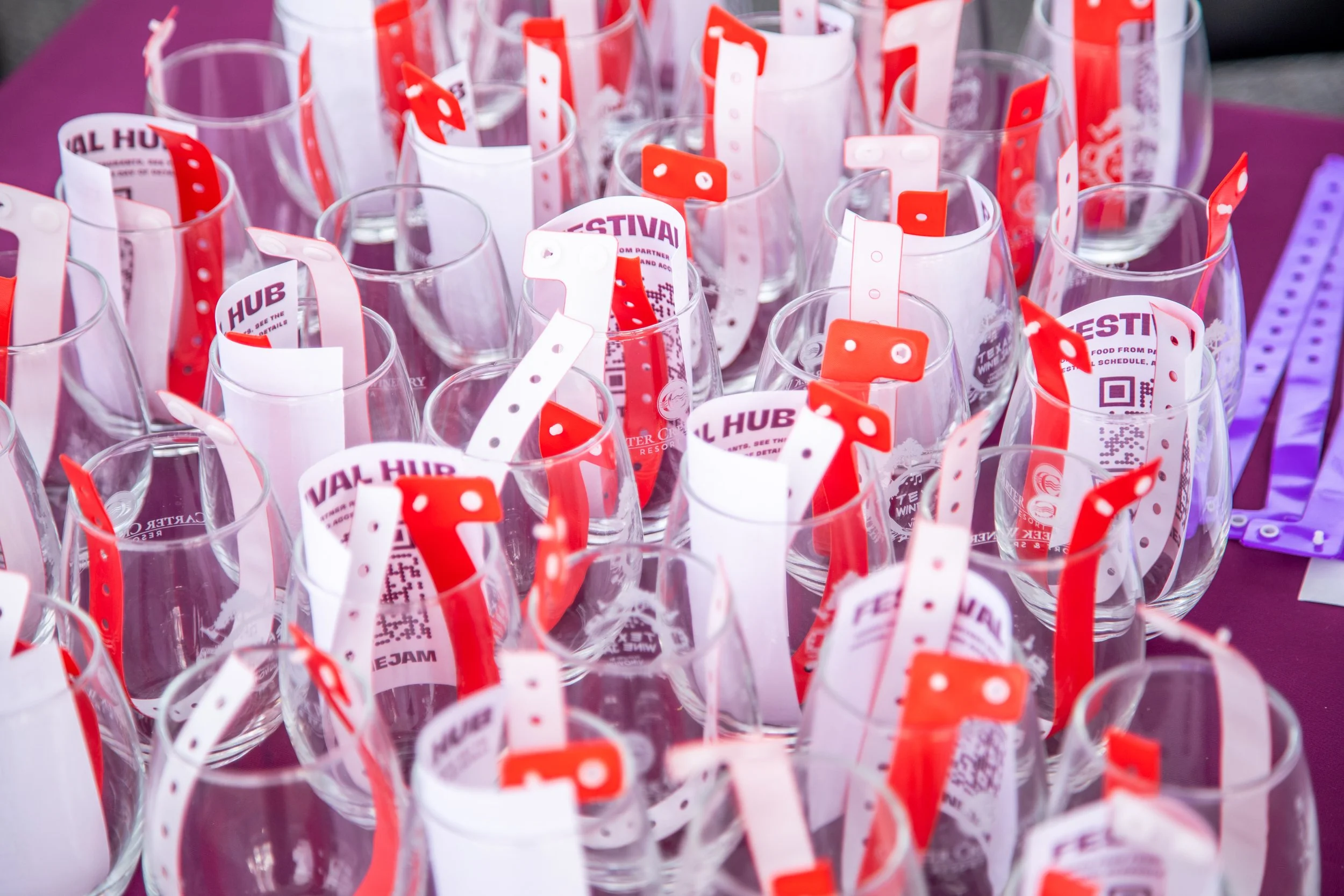 Collection of clear glass cups with red and white event tickets inside, placed on a purple tablecloth.