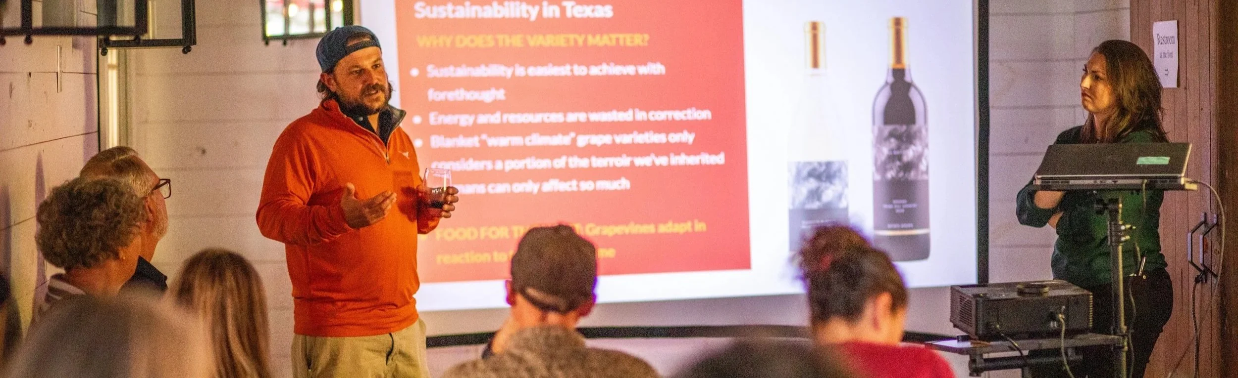 A man in an orange jacket and a backward baseball cap is giving a presentation on sustainability in Texas to an audience. He is holding a glass of wine and gesturing with his other hand. A woman on the right is standing near a projector and laptop, listening. The presentation slide behind them discusses the importance of sustainability and related topics.