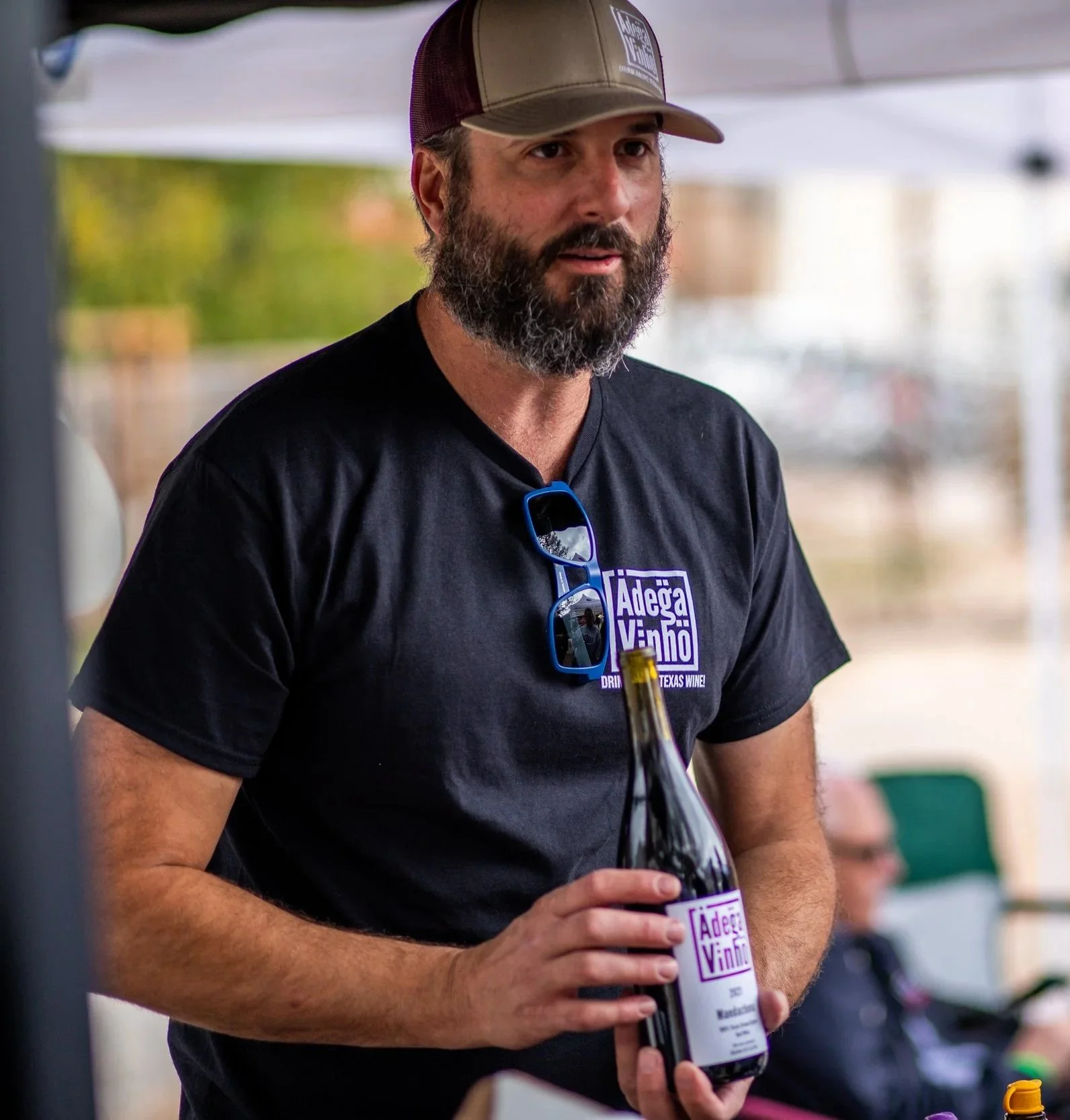 Man with a beard wearing a brown and black trucker hat and black t-shirt with a purple logo, holding a beer bottle with a white label, in an outdoor setting.